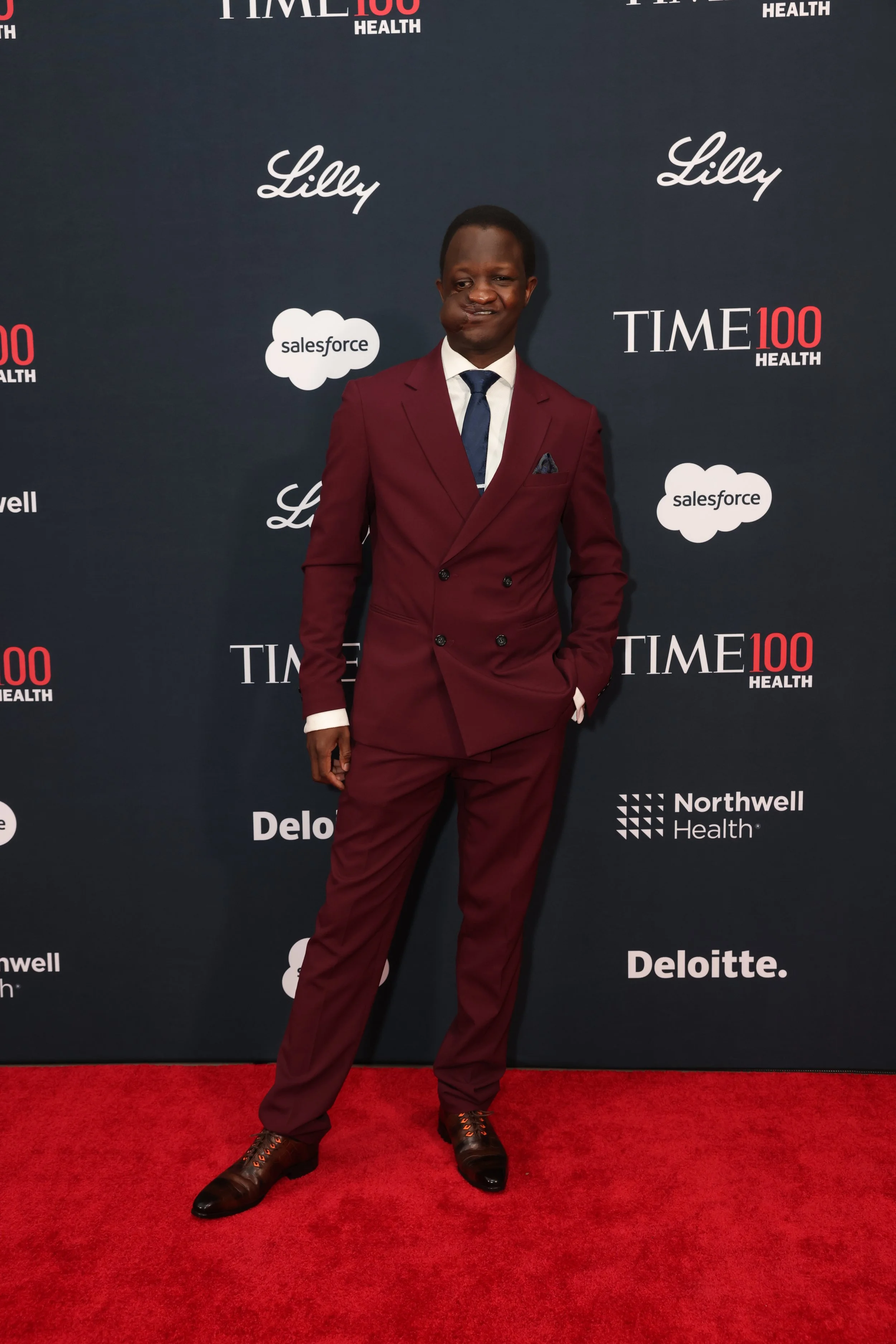A man in a burgundy suit with a white shirt and dark blue tie, standing on a red carpet at a Time 100 Health event, posing in front of a navy blue backdrop with logos and text including Salesforce, Lilly, and Deloitte.