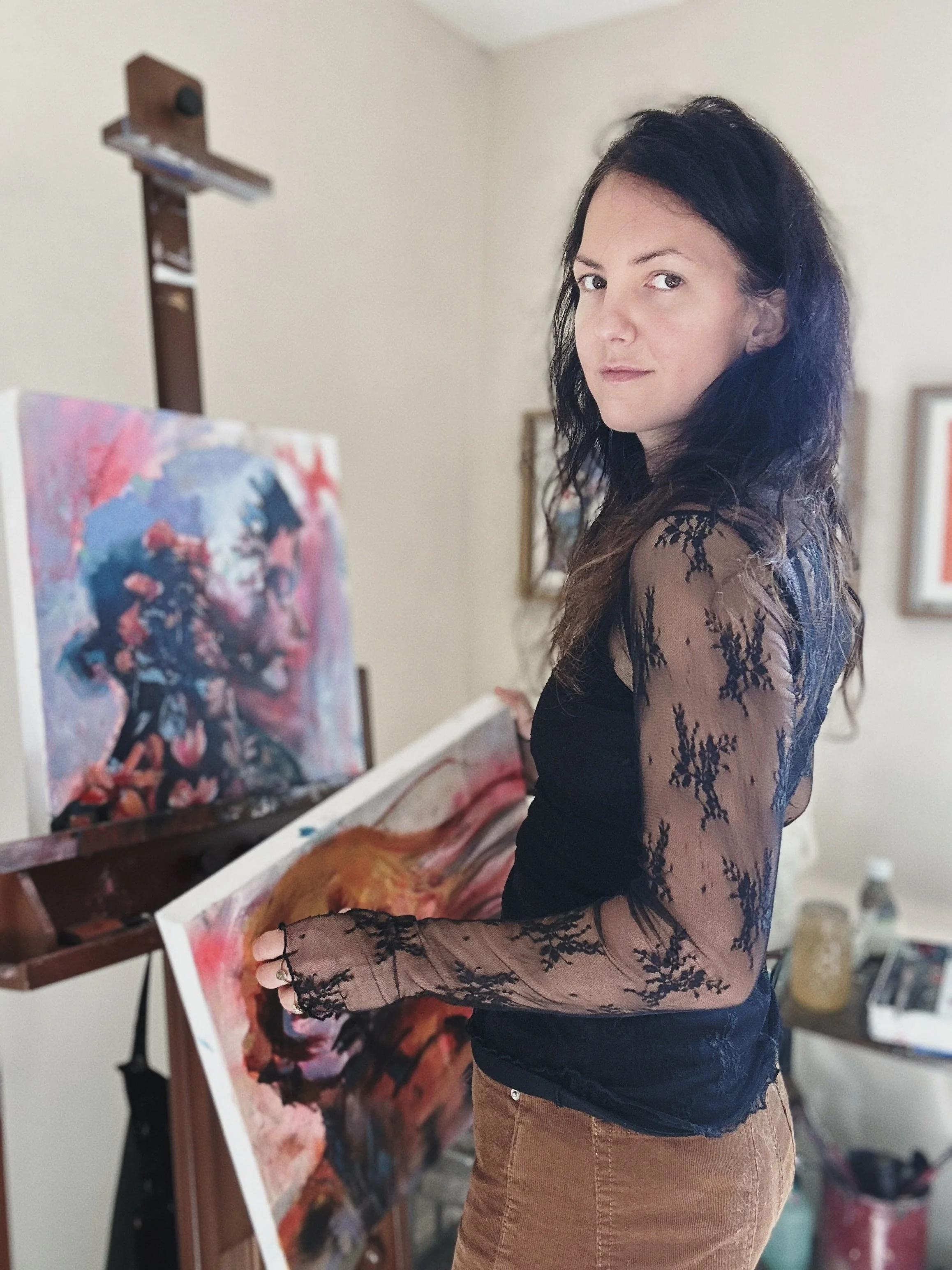 A woman with long dark hair, wearing a black lace long sleeve top and brown pants, stands next to an easel with a colorful abstract painting in progress, holding a canvas in an art studio.