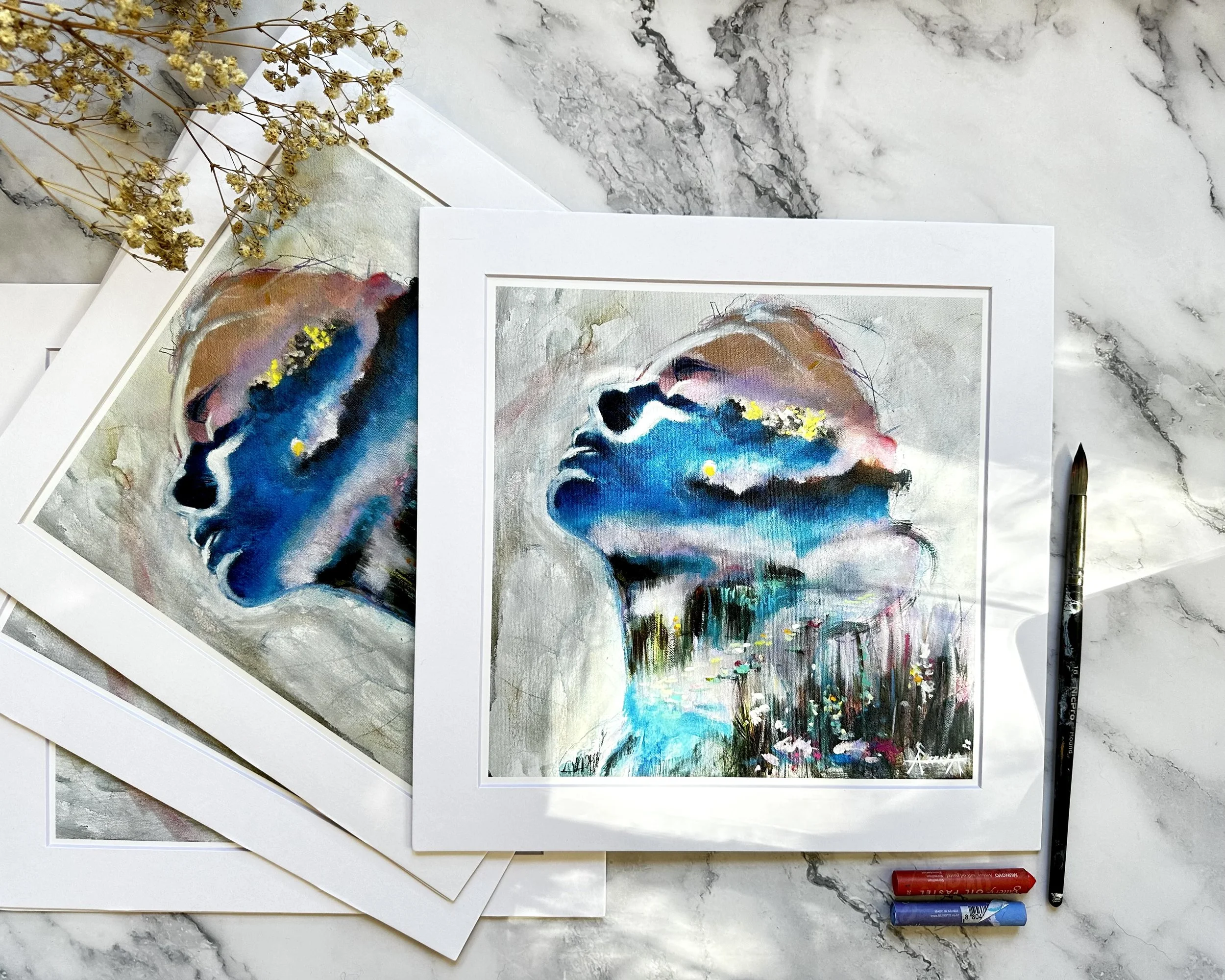 Hand embellished, affordable, signed original fine art prints of a woman's face with closed eyes, featuring vibrant blues, pinks, yellows, and abstract elements, laid out on a marble surface with art supplies and dried flowers nearby.