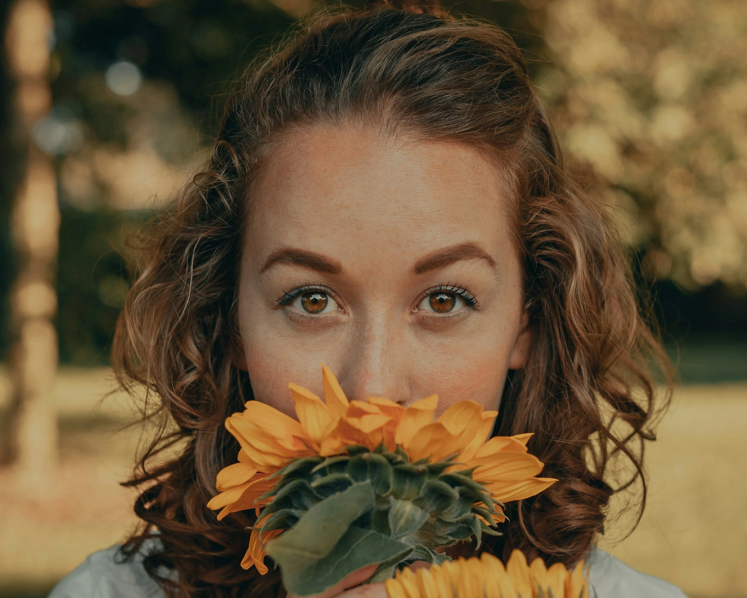 A woman with curly brown hair and brown eyes holding a bouquet of orange sunflowers in front of her face outdoors.