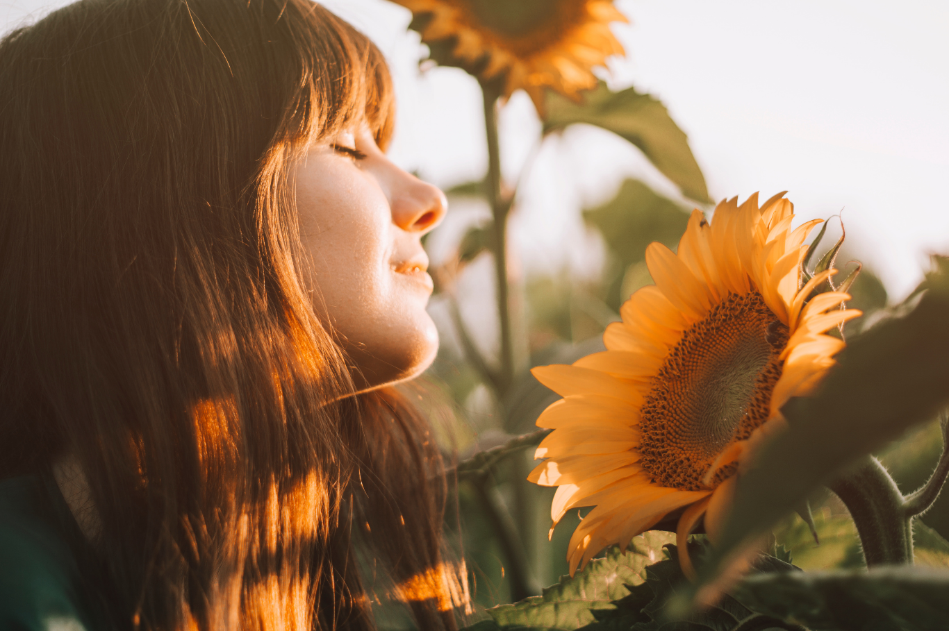 A woman with red hair stands close to a sunflower during sunset, with her eyes closed and a peaceful expression.