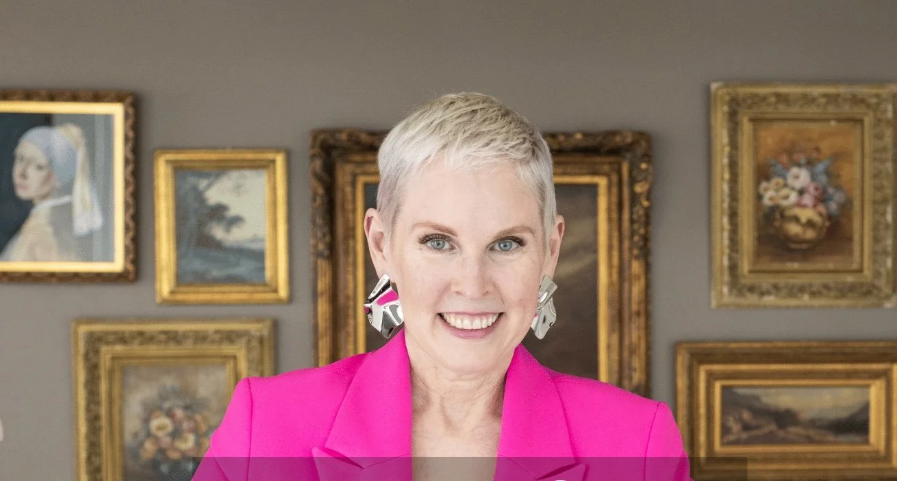A woman with short blonde hair, wearing a bright pink blazer and large abstract earrings, smiling in front of a gallery wall with framed paintings.
