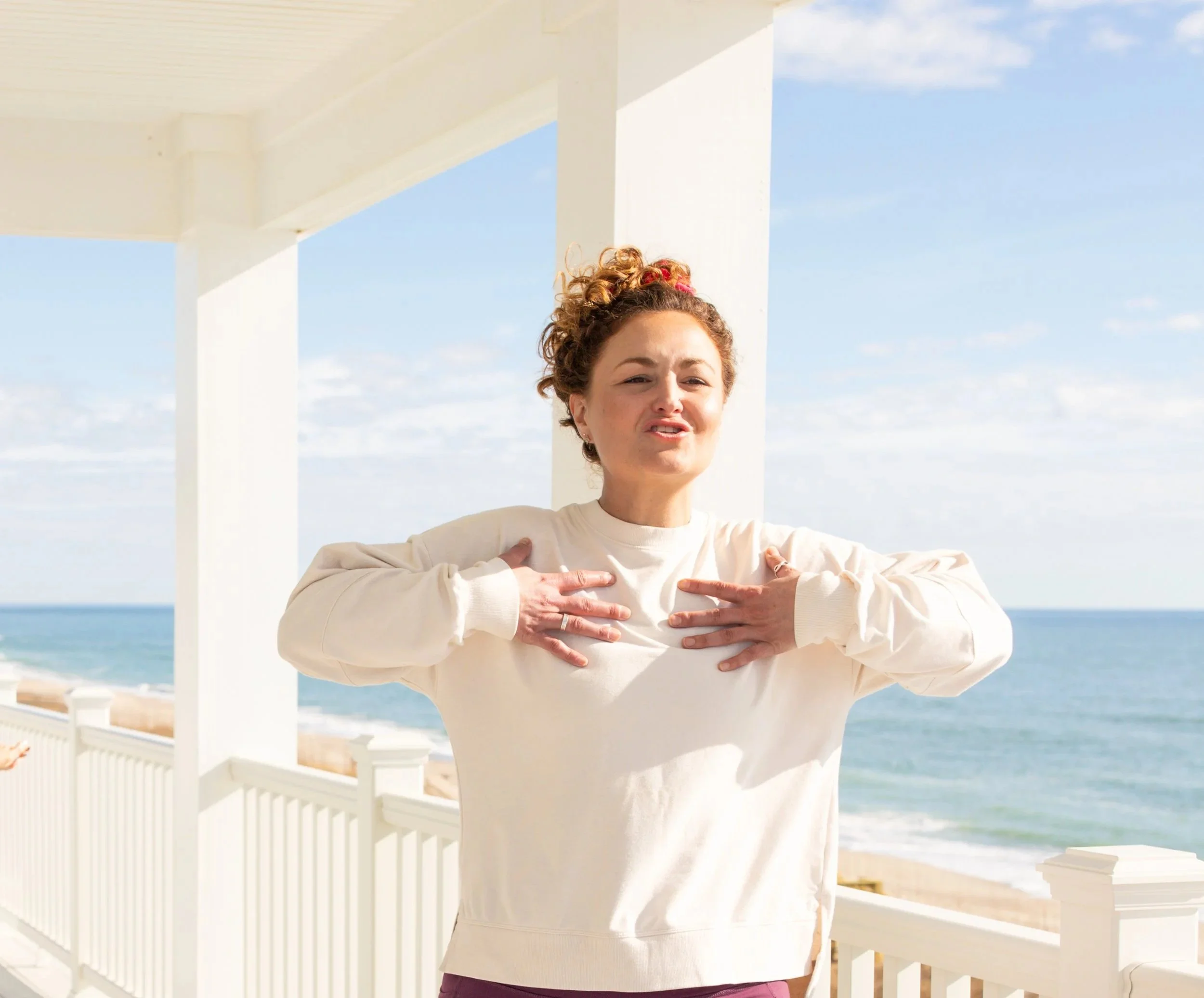 A woman with curly hair and light skin standing on a balcony near the beach, with her hands on her chest, making a funny face.