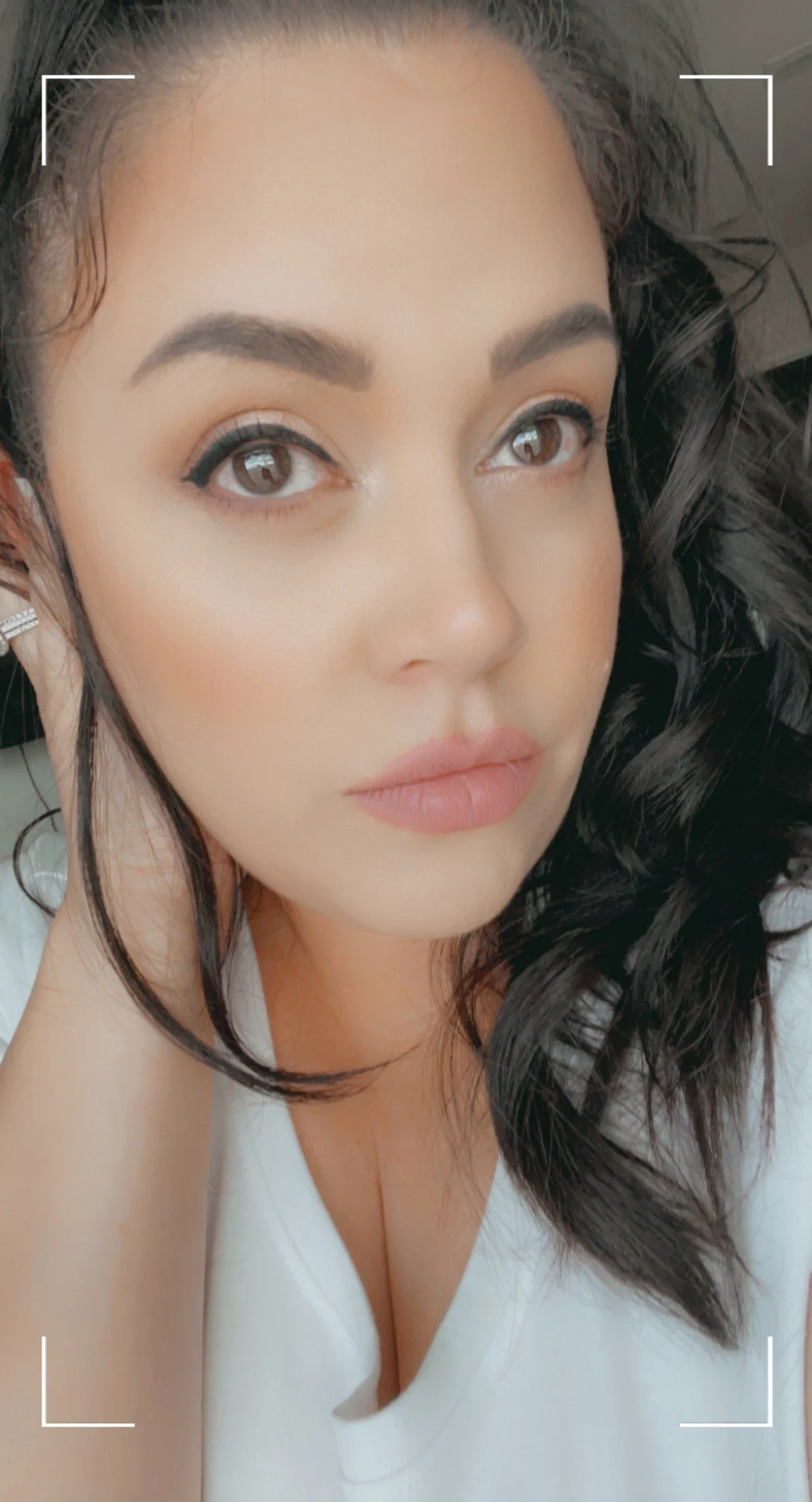 Close-up of a woman with dark wavy hair, light makeup, and wearing a white top, resting her head on her hand.