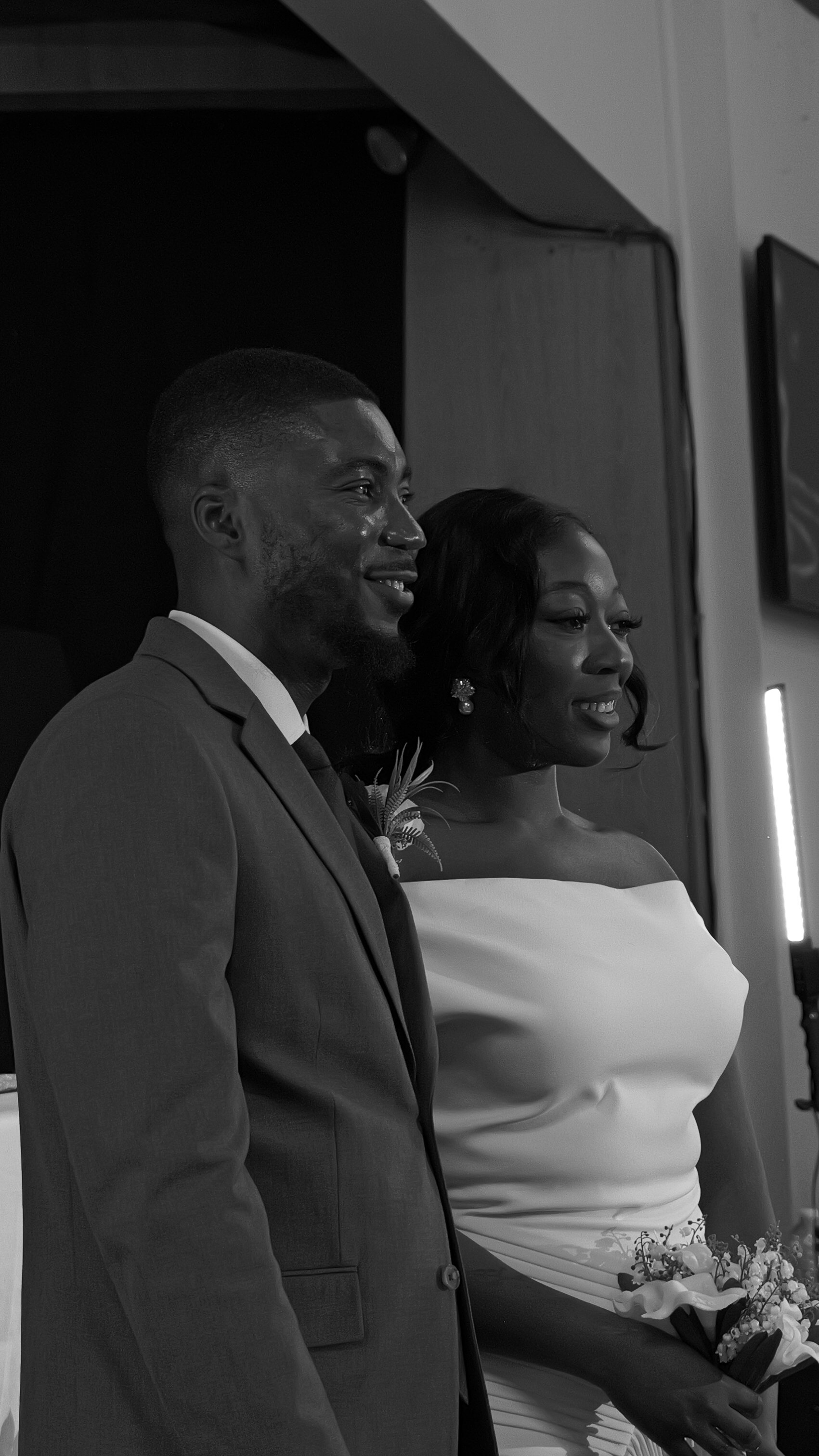 A woman with long, wavy black hair is smiling and looking to the side in a dimly lit room with purple lighting, wearing a beige off-shoulder top.