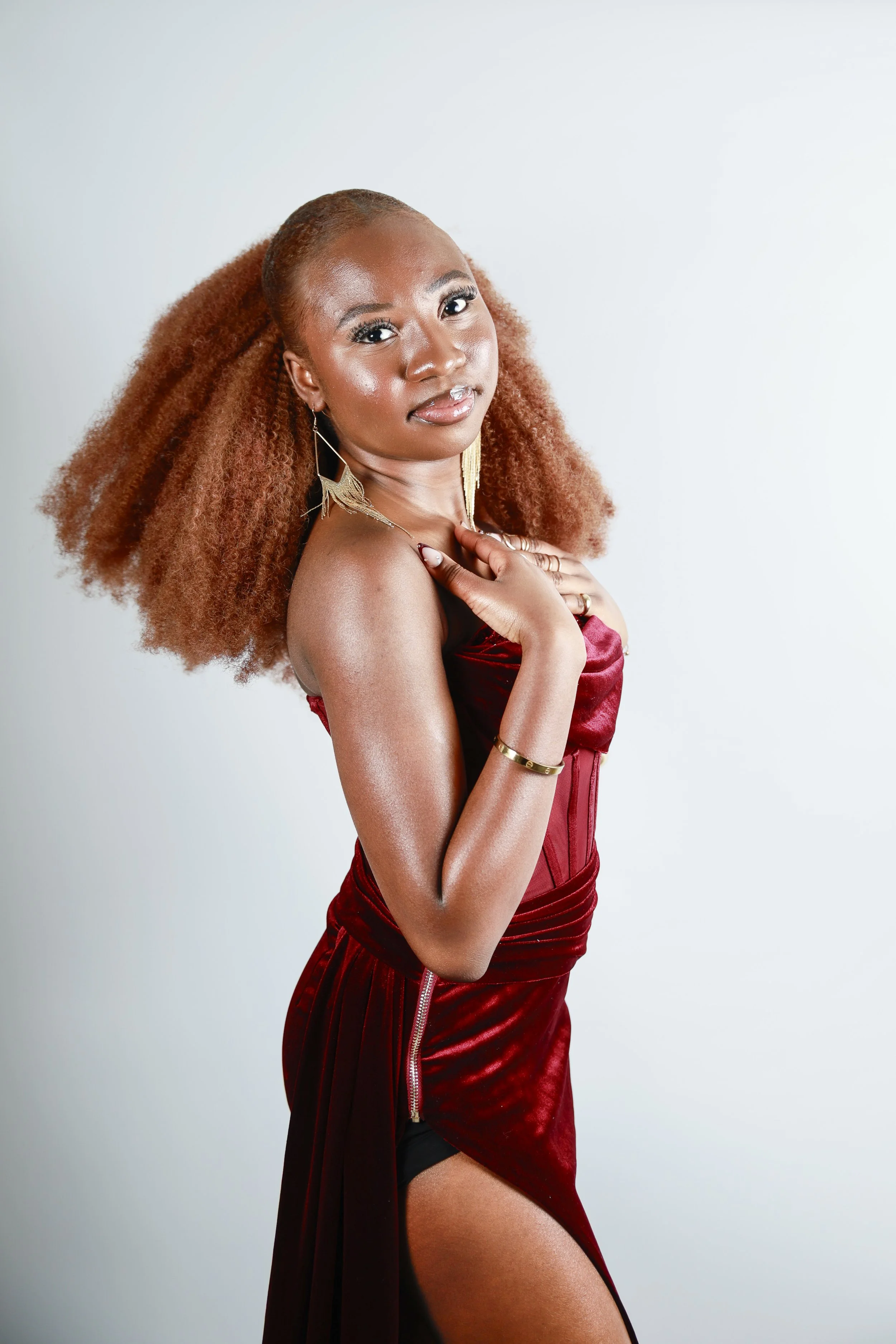 A woman with dark skin dressed in traditional African attire, wearing a patterned dress and headwrap in shades of blue, with red jewelry, standing against a brown background.