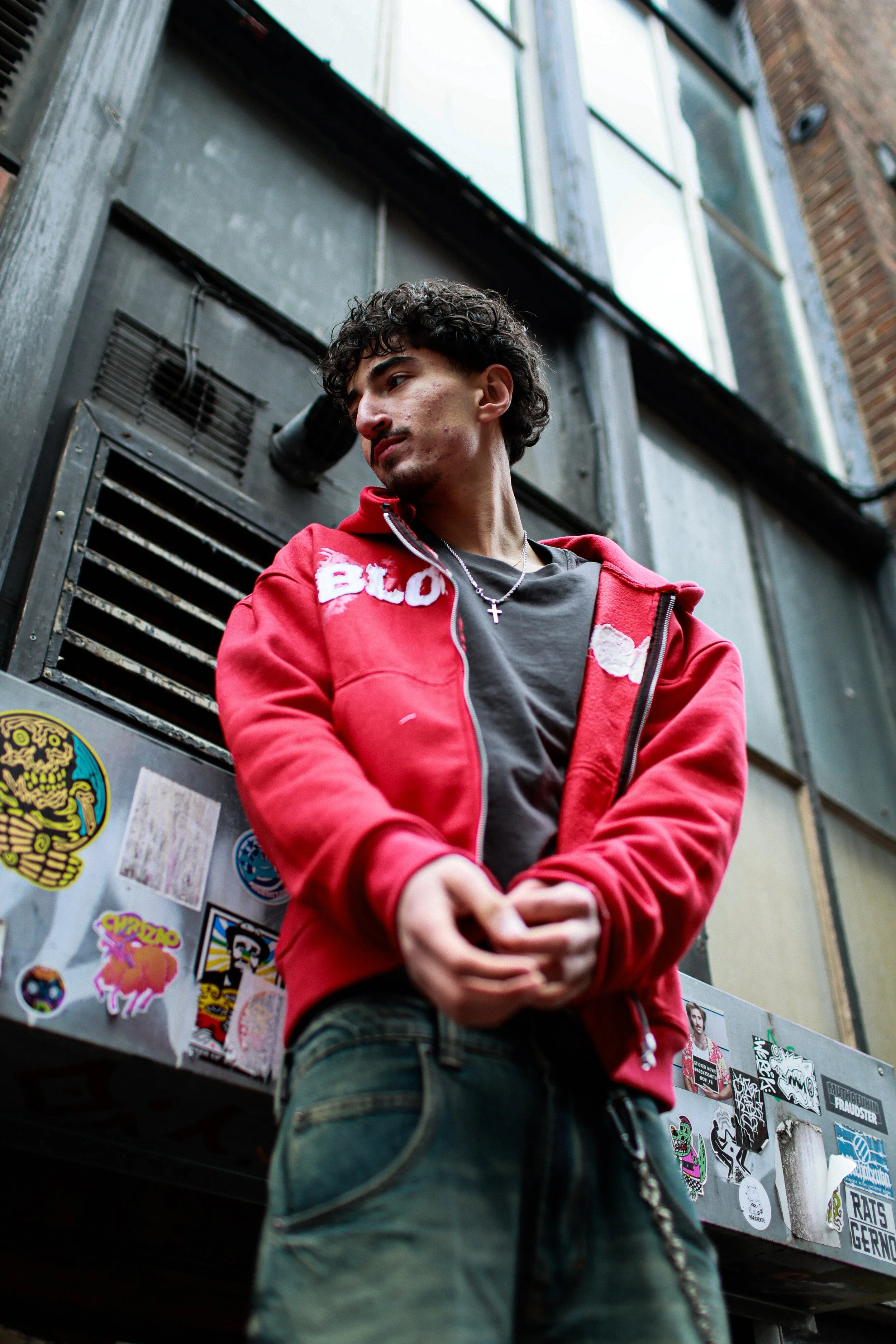 A person playing an electric bass guitar on stage, wearing a striped shirt, a cap, and jewelry, with a dark background.