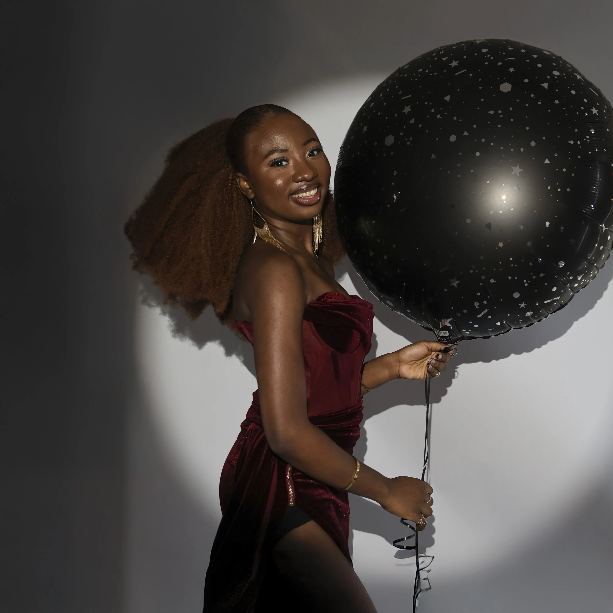Two women smiling and looking at each other, wearing matching cream-colored dresses with gold sequins, standing against a brown background.