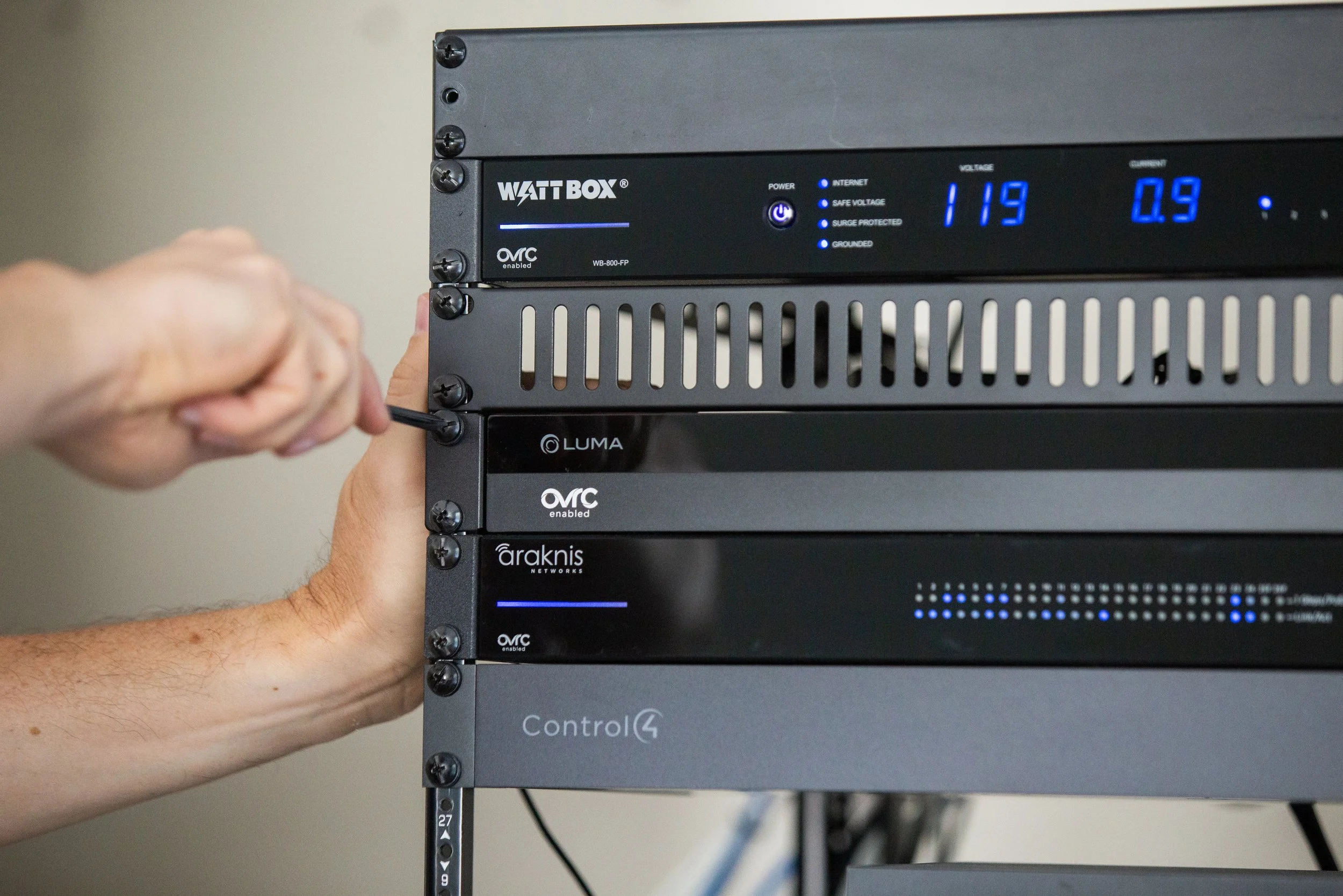 A person using a screwdriver to tighten a screw on a black server rack with multiple network devices and a blue digital display showing voltages and currents.