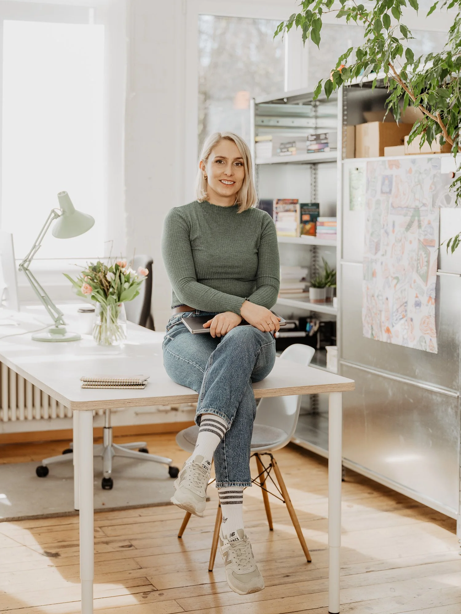 Eine junge Frau sitzt lächelnd auf einem Schreibtisch in einem hellen Büro. Sie trägt eine grüne Strickpullover, Jeans, weiße Sportschuhe und Socken. Im Vordergrund ein Computer, auf dem Schreibtisch eine Vase mit Blumen, ein Schreibtischlampe und Notizbuch. Im Hintergrund Bücherregale, Pflanzen und ein Whiteboard mit Zeichnungen.