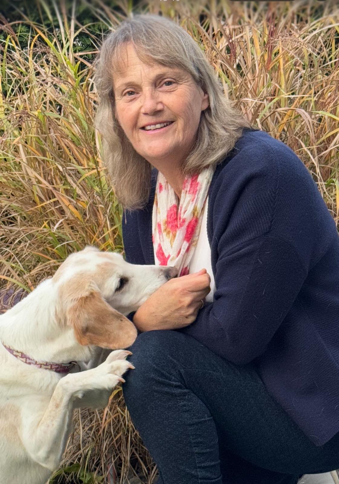 Eine Frau sitzt im Feld mit hohen Gräsern und hält einen Welpen im Arm, der sich an sie kuschelt.