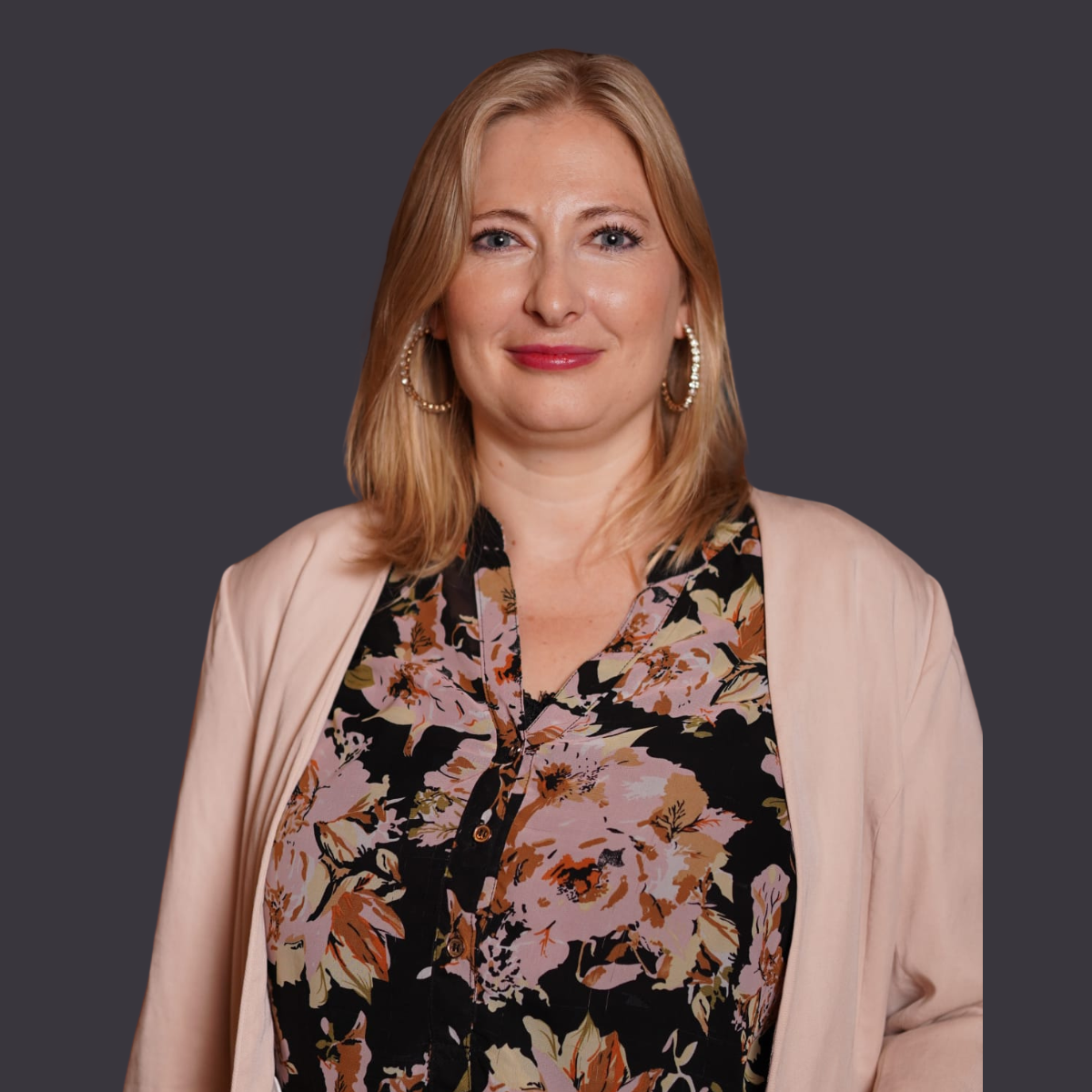 Porträt einer Frau mit blonden Haaren, blauer Augen, roten Lippen und großen goldenen Ohrringen, trägt ein blumiges Hemd und eine beige Jacke vor dunklem Hintergrund.