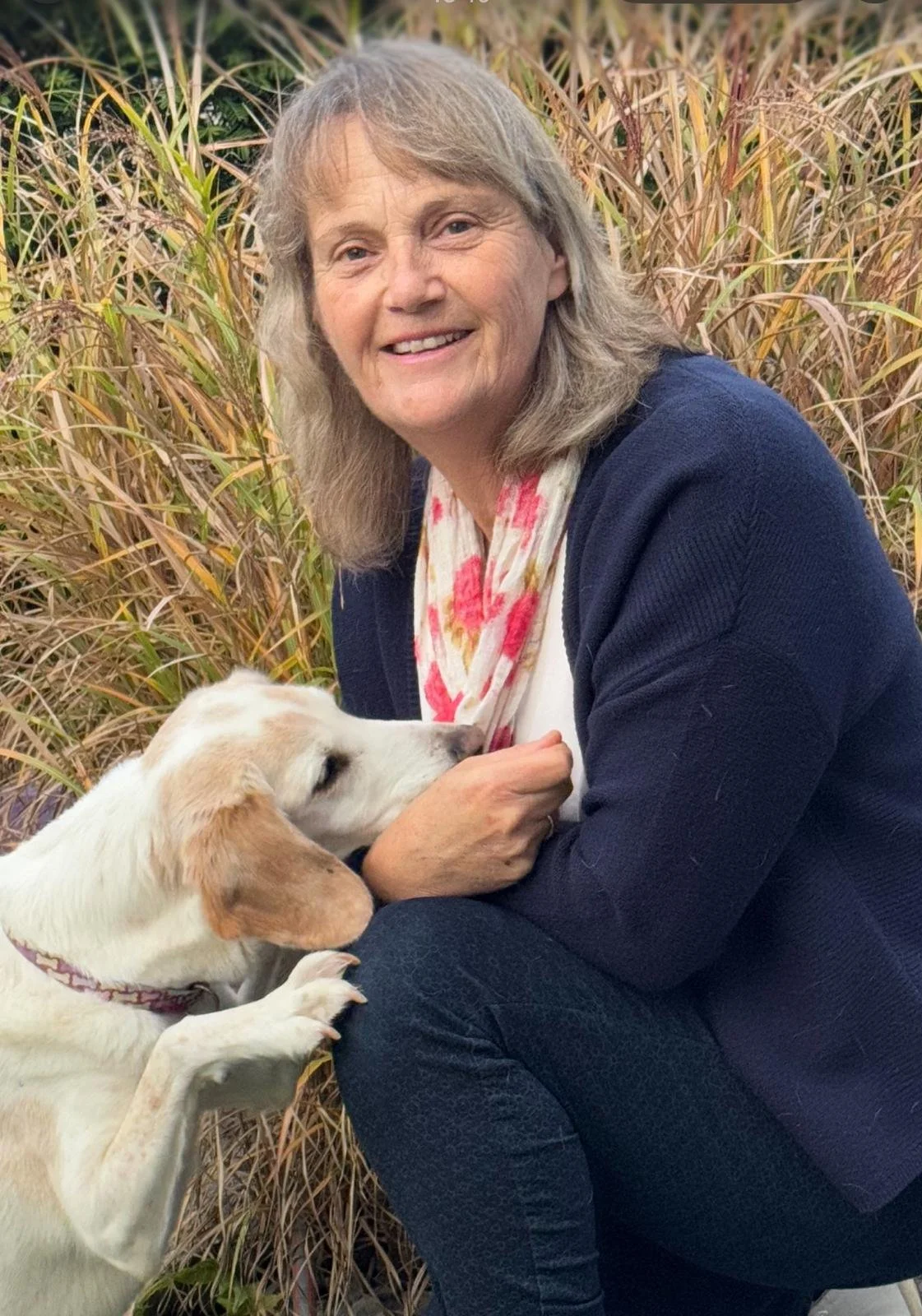 Eine ältere Frau sitzt in einem Feld mit Gräsern, lächelt und hat einen weißen Hund mit braunen Ohren, der an ihrer Hand leckt.