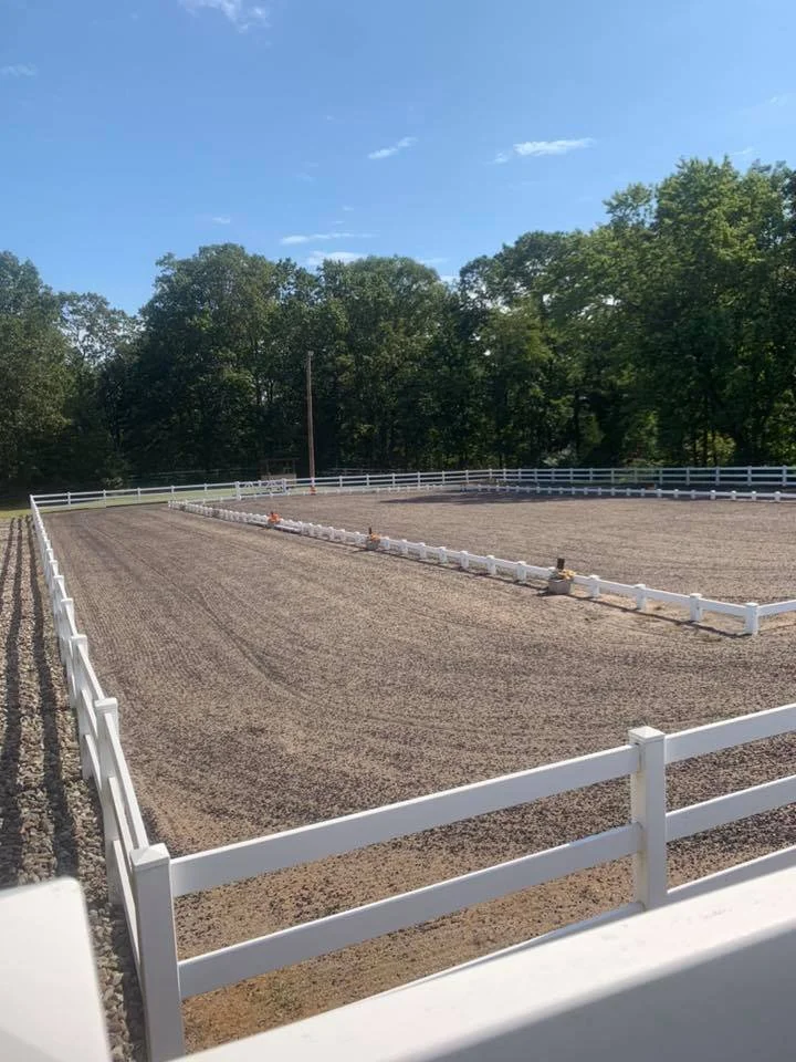 Empty horse riding arena with white fencing and trees in the background under a clear blue sky.