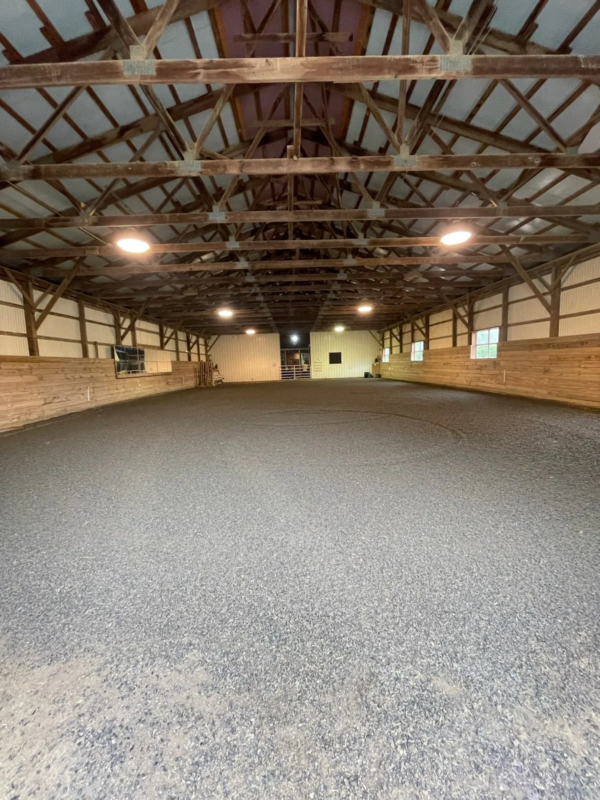 Empty indoor riding arena with rubber-sand footing, wooden walls, and a high ceiling with exposed wooden beams and overhead lighting.