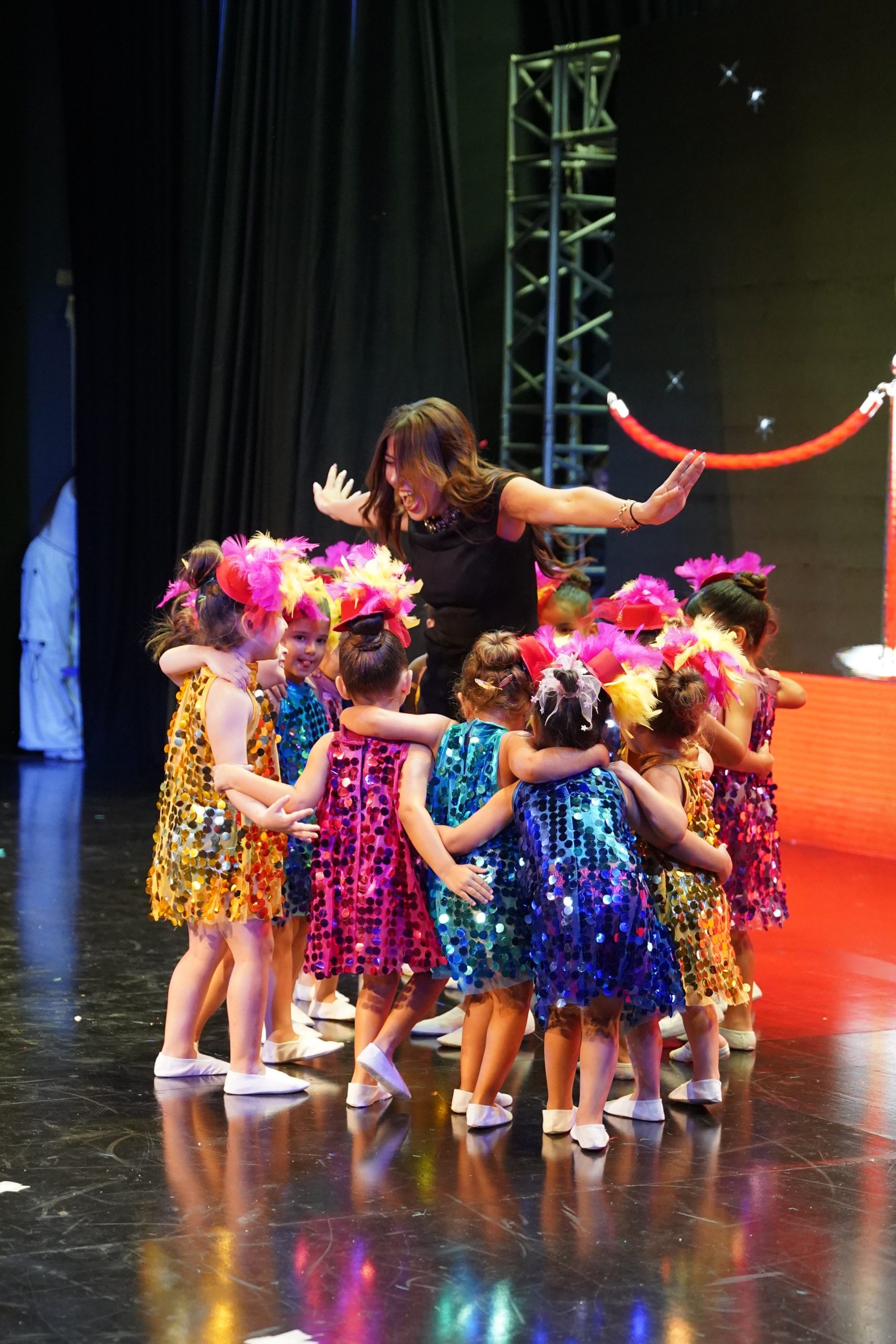A group of young girls in colorful sequin dresses are hugging and celebrating with a woman on a stage during a performance, with stage lighting and a dark background.