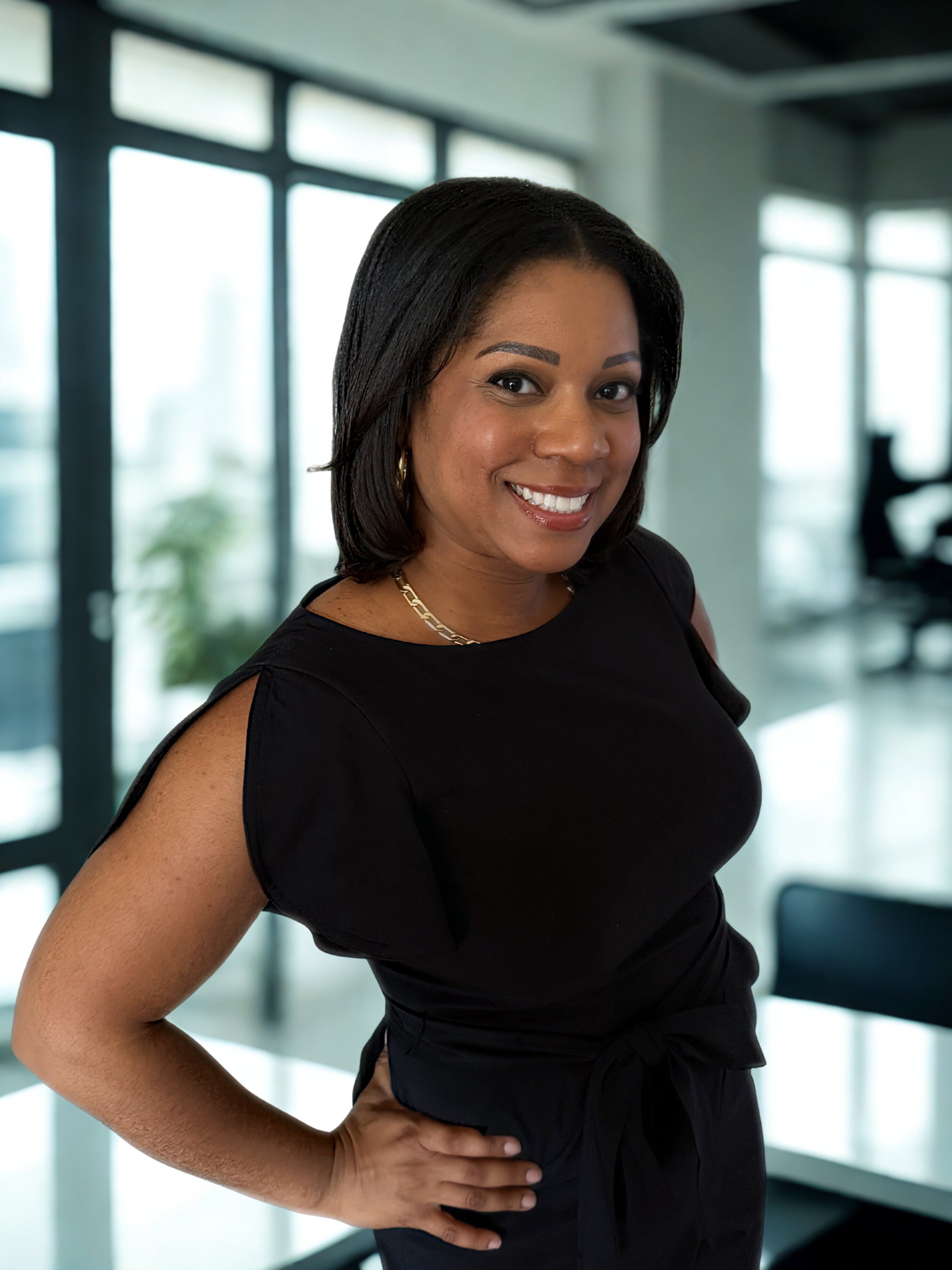 Smiling woman in black dress posing in modern office building with large windows.