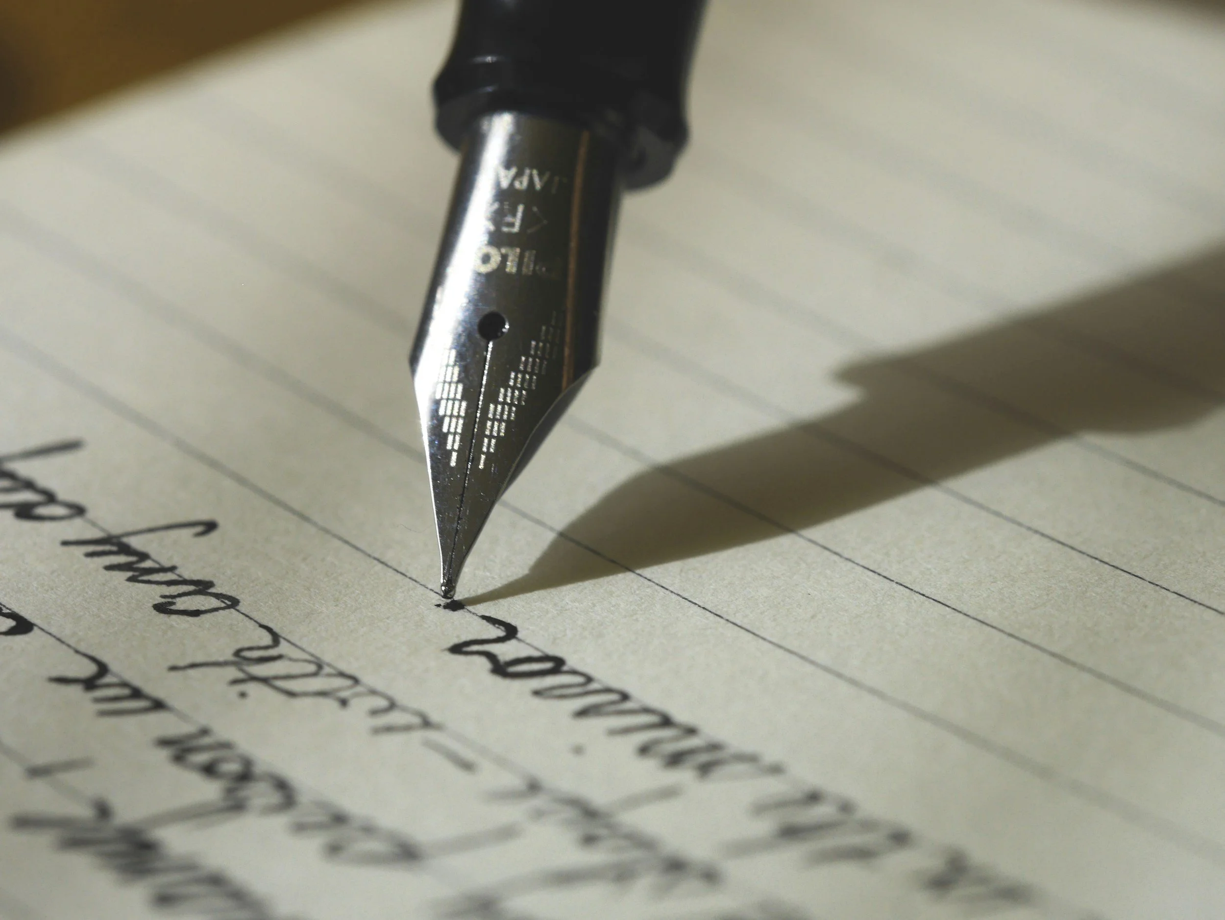 Close-up of a fountain pen writing on lined paper with black ink, with the pen's shadow visible on the paper.