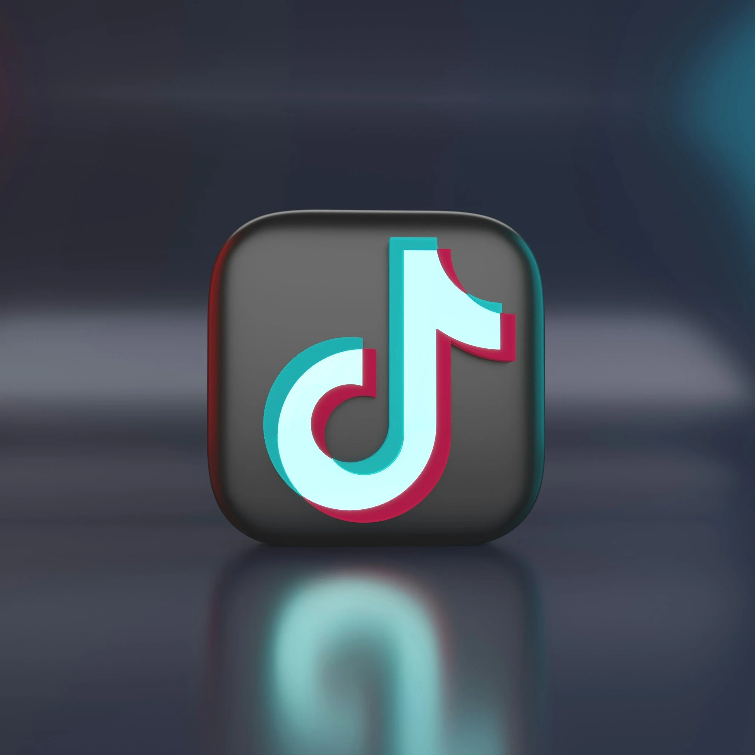 TikTok logo on a gray 3D app icon with reflective surface and blurred dark background.