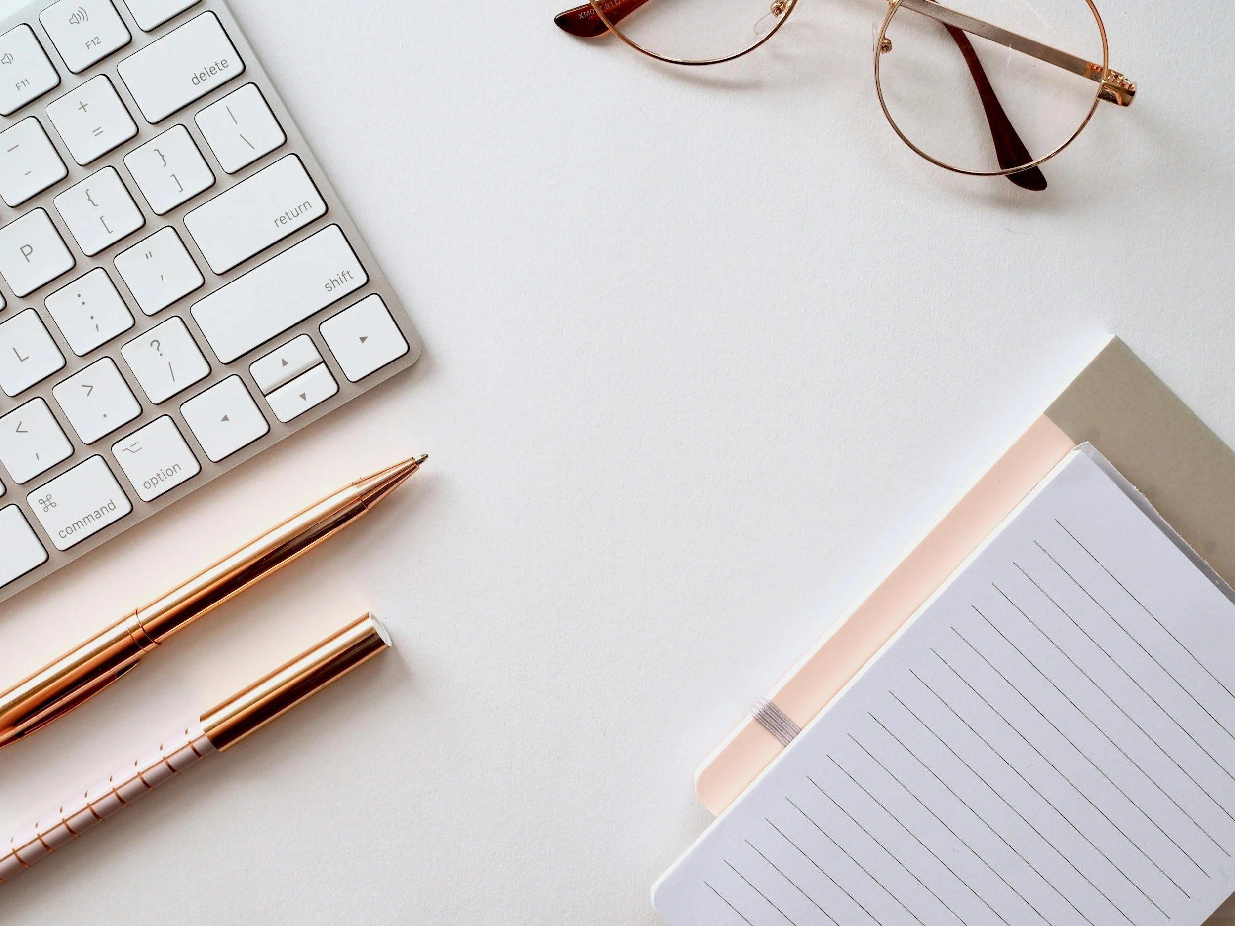 A white desk with a computer keyboard, a pair of sunglasses, two pens, and an open notebook with lined pages.