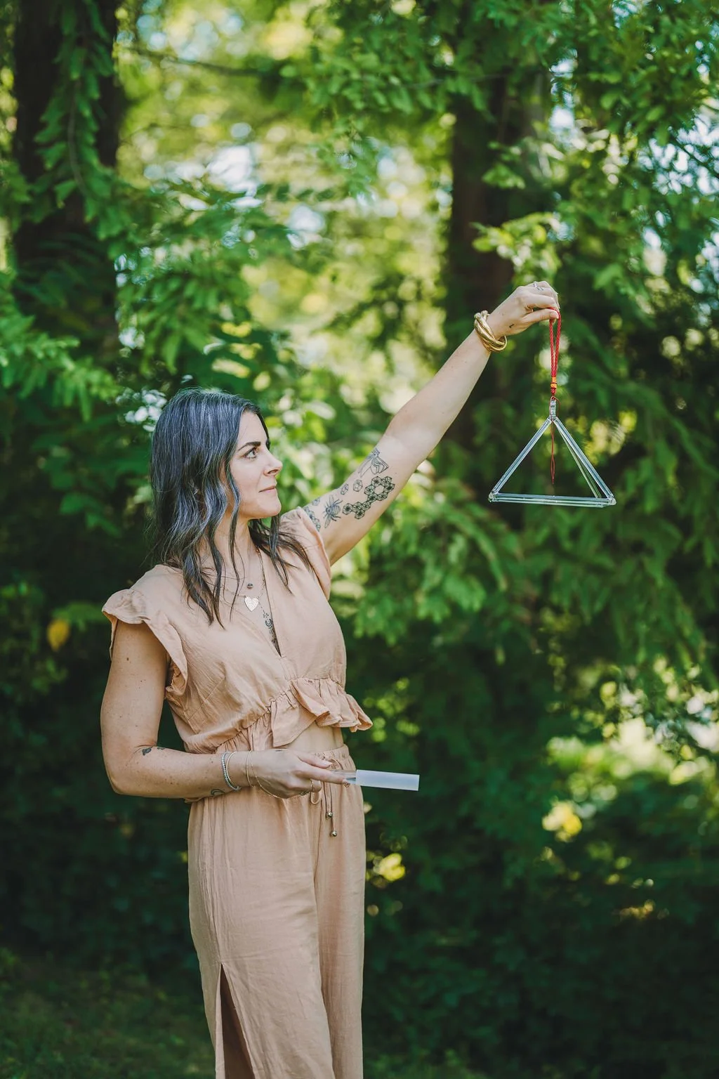 A woman with dark hair and tattoos, wearing a beige outfit, stands outdoors surrounded by green trees. She is holding a triangle-shaped pendulum attached to a red cord, with her arm extended upward, while holding a small white object in her other hand.