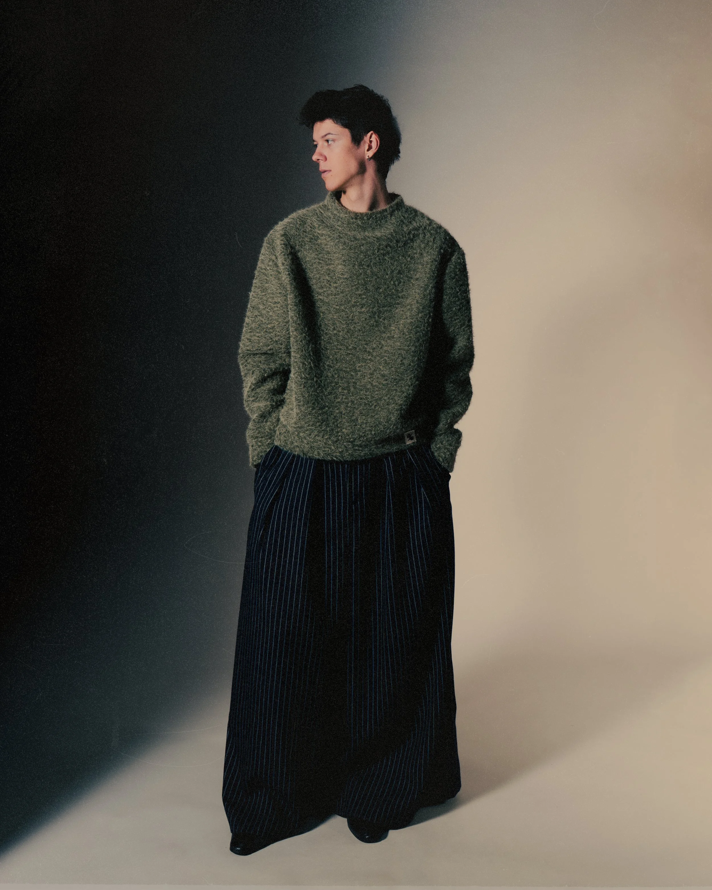 Une personne avec des cheveux courts et noirs, portant un pull vert foncé et un pantalon large à rayures bleues, se tient contre un fond en dégradé du noir au beige.