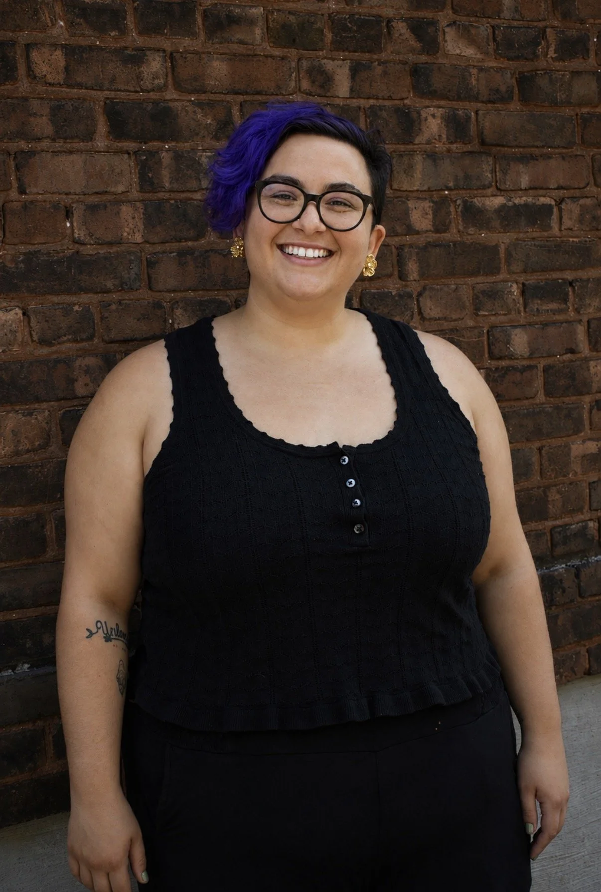 A woman with short, purple and black curly hair smiling outdoors while wearing a black sleeveless top with gold earrings.