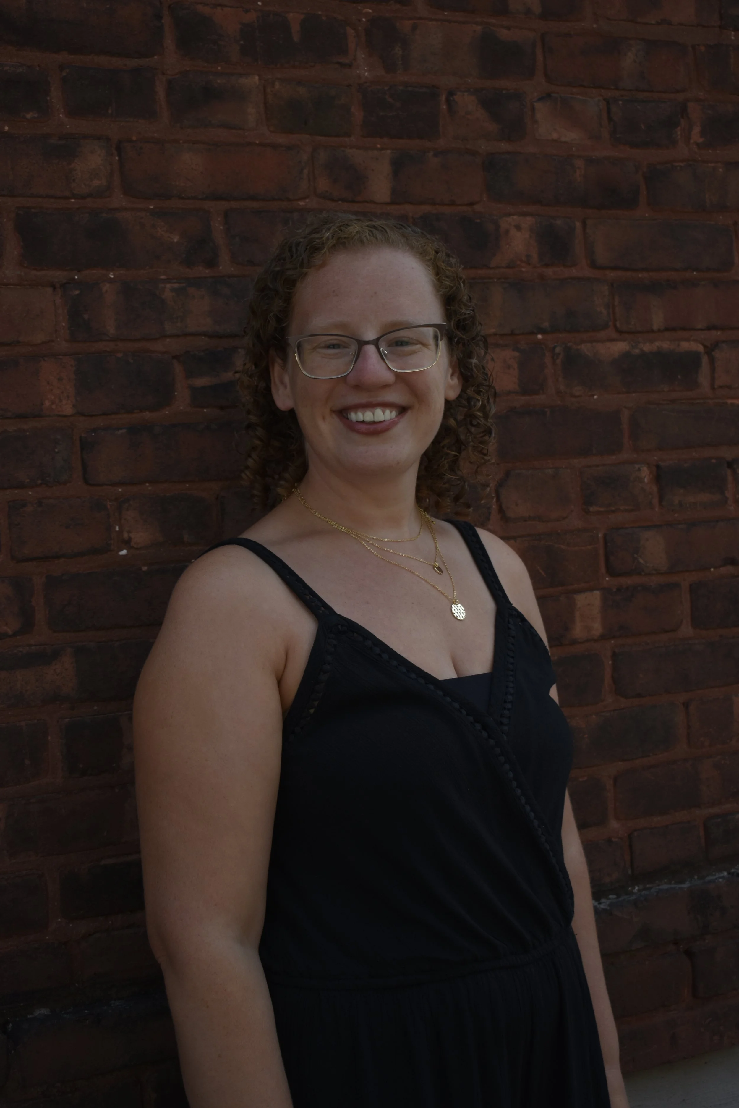 A woman with curly red hair, glasses, and a striped tank top is smiling and looking up outdoors near a tree on a sunny day.