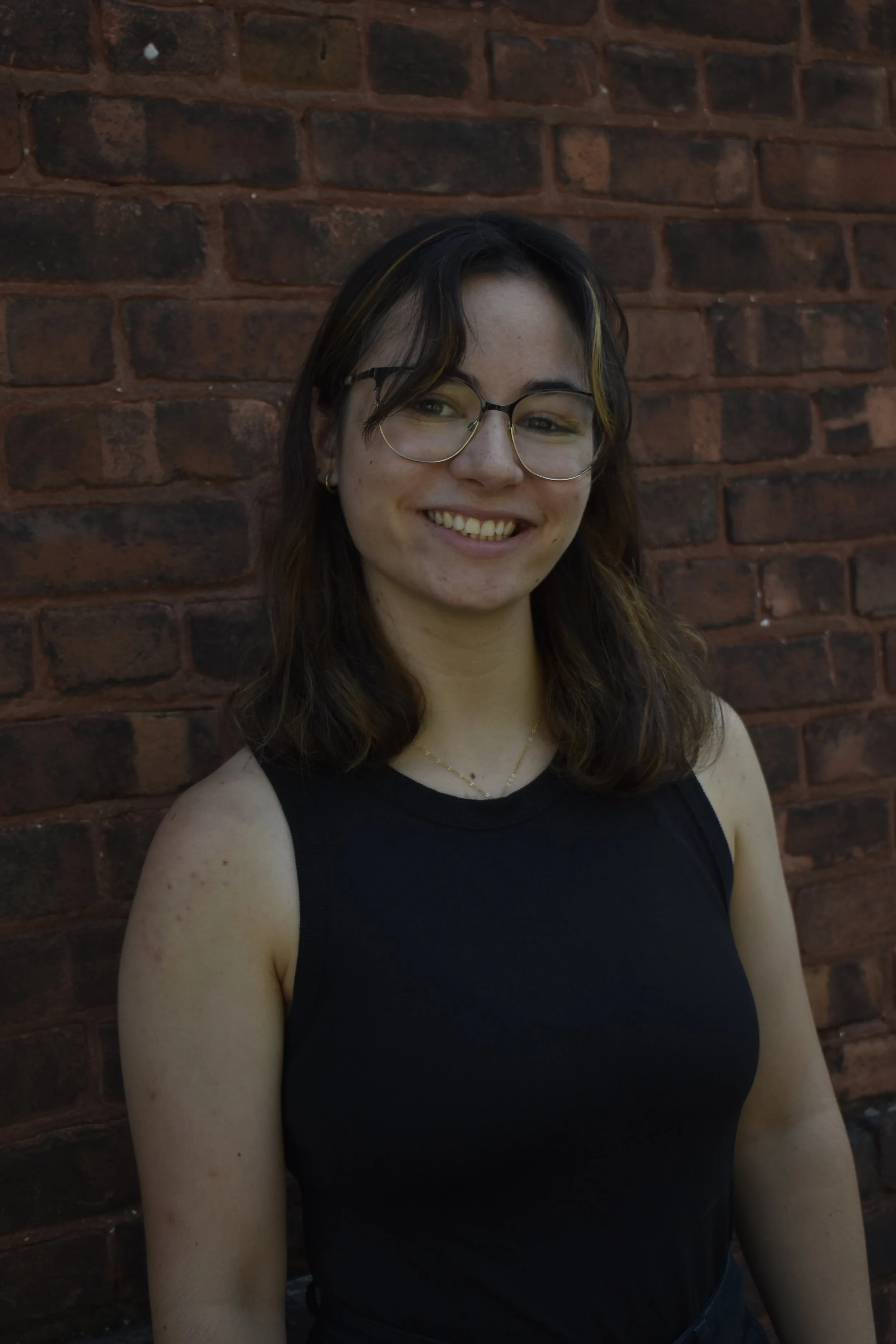 A young woman with dark brown hair and fair skin smiling, wearing a white sleeveless top, in front of a brick wall.