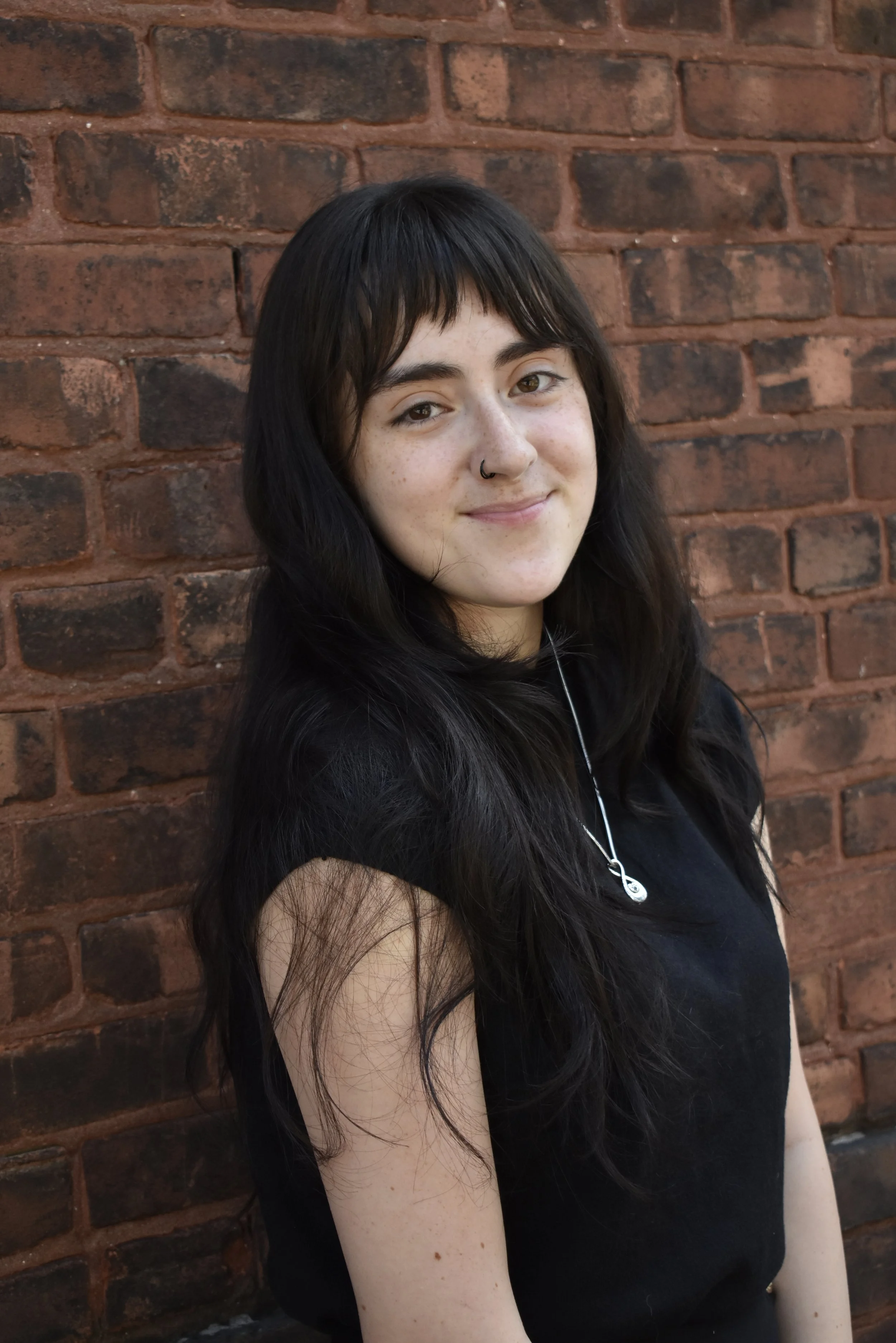 Close-up photo of a young woman with long dark hair, wearing a nose ring, smiling softly outdoors with a background of green trees.