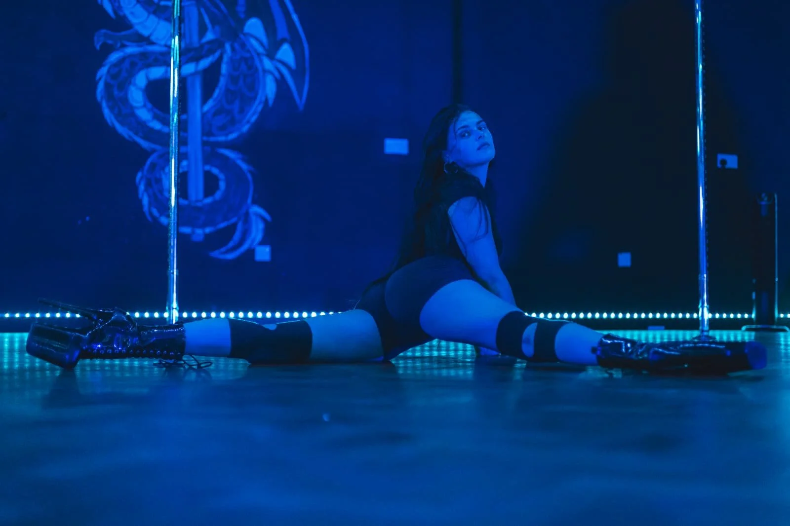 A woman performs split on the floor of a dance studio illuminated with blue lighting, with a neon snake artwork on the wall behind her.