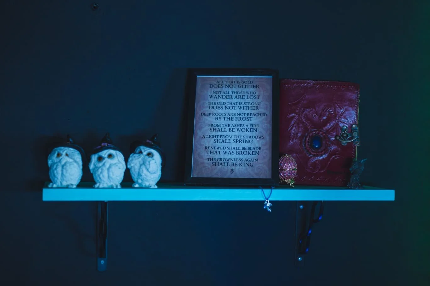 A blue floating shelf with three owl figurines and decorative items, a framed poem, and a red book with dragon embroidery, set against a dark wall.