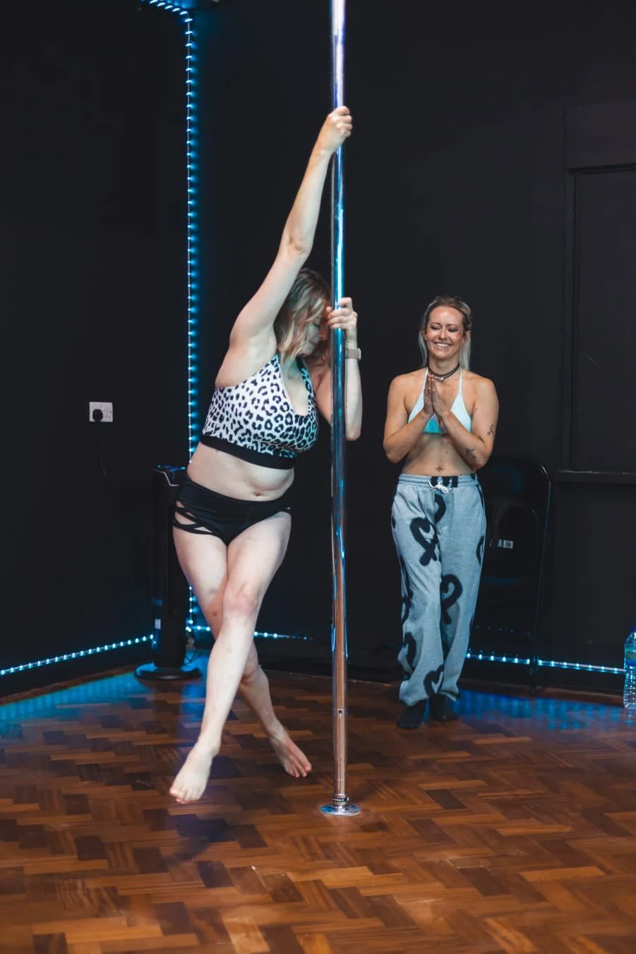 A woman performing a pole dance routine with an instructor observing happily in a dance studio.