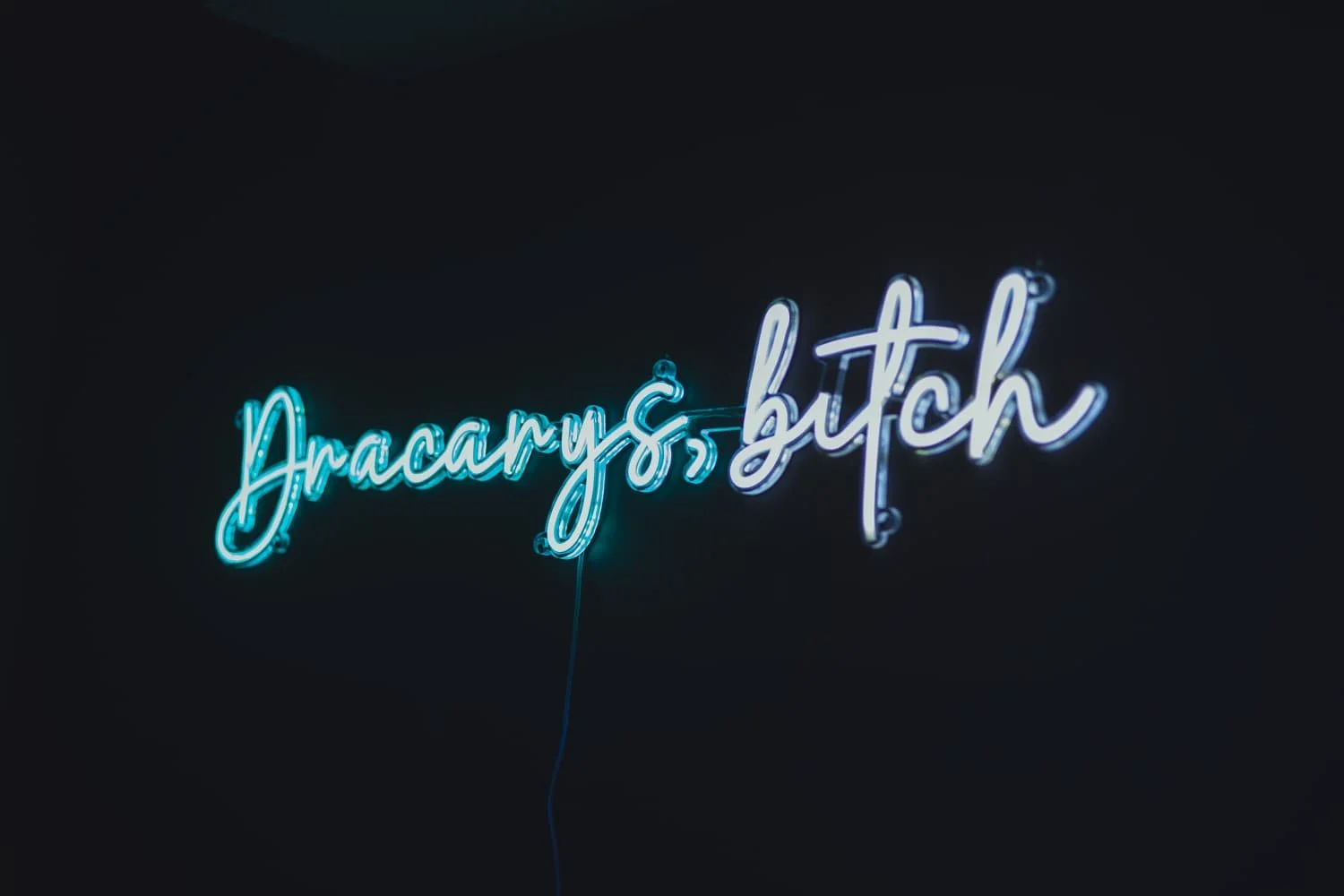 Neon sign that reads 'dracarys, bitch' in cursive blue and white letters against a dark background.