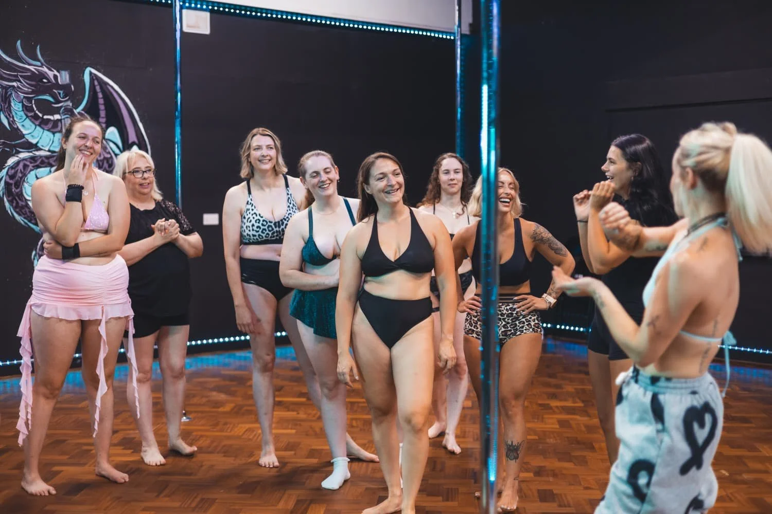 A group of women in swimwear listening to a woman in a sports bra and shorts talking and gesturing during a dance class in a studio with dark walls and decorative lighting.