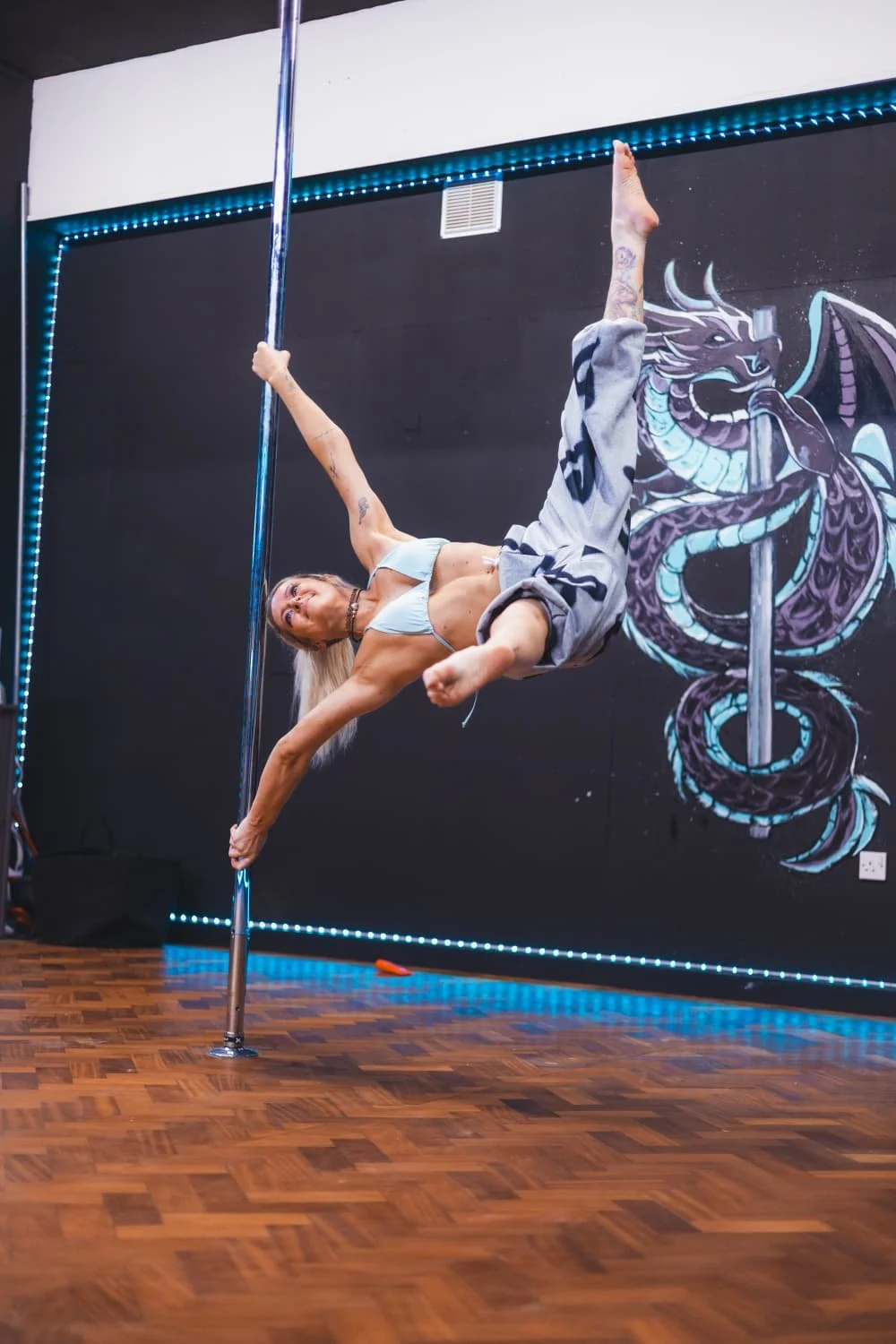 A woman performing pole dance moves on a vertical pole in a studio with dark walls and a dragon mural.