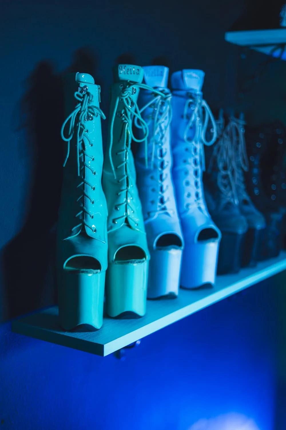 A row of blue, lace-up, open-toe platform high heels on a shelf, dimly lit with blue lighting.