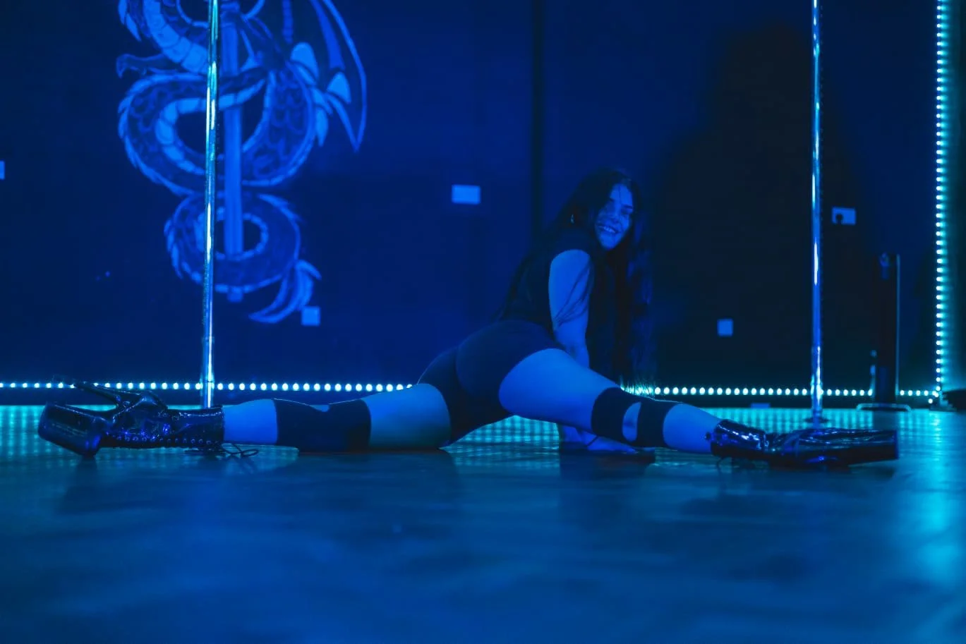 Woman stretching on the floor of a dance studio with blue neon lighting and a dragon mural on the wall.