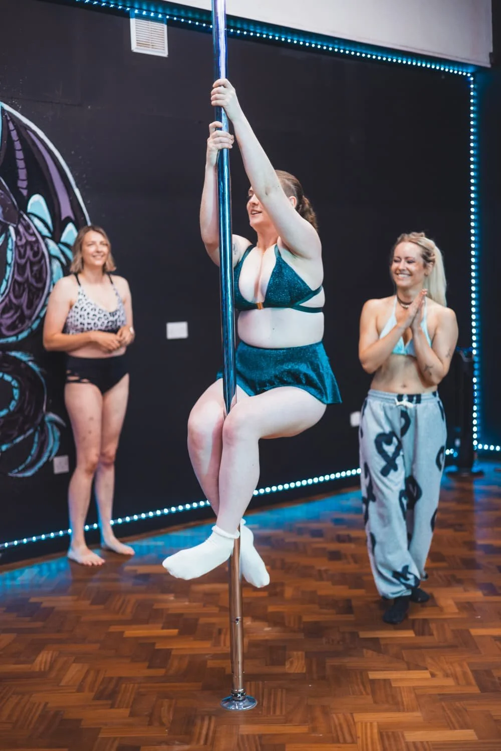 A woman performing pole dance moves while three women watch and cheer in an indoor studio with black walls and blue lighting.