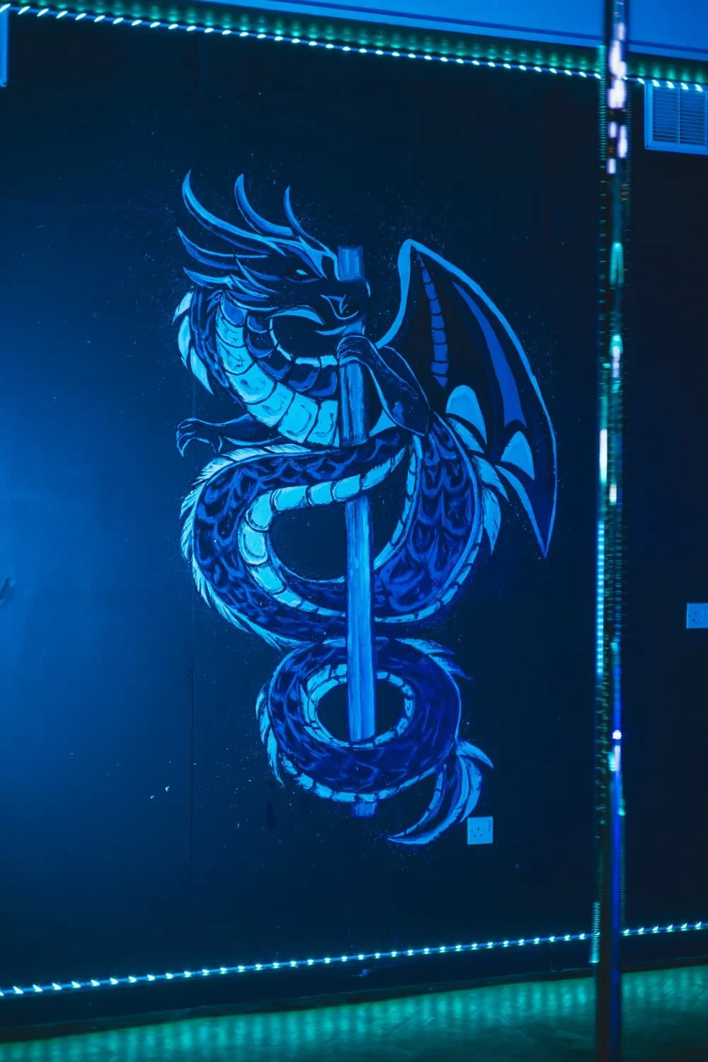 Neon-lit artwork of a dragon with wings coiled around a pole, illuminated in blue with light strips on the wall edges.