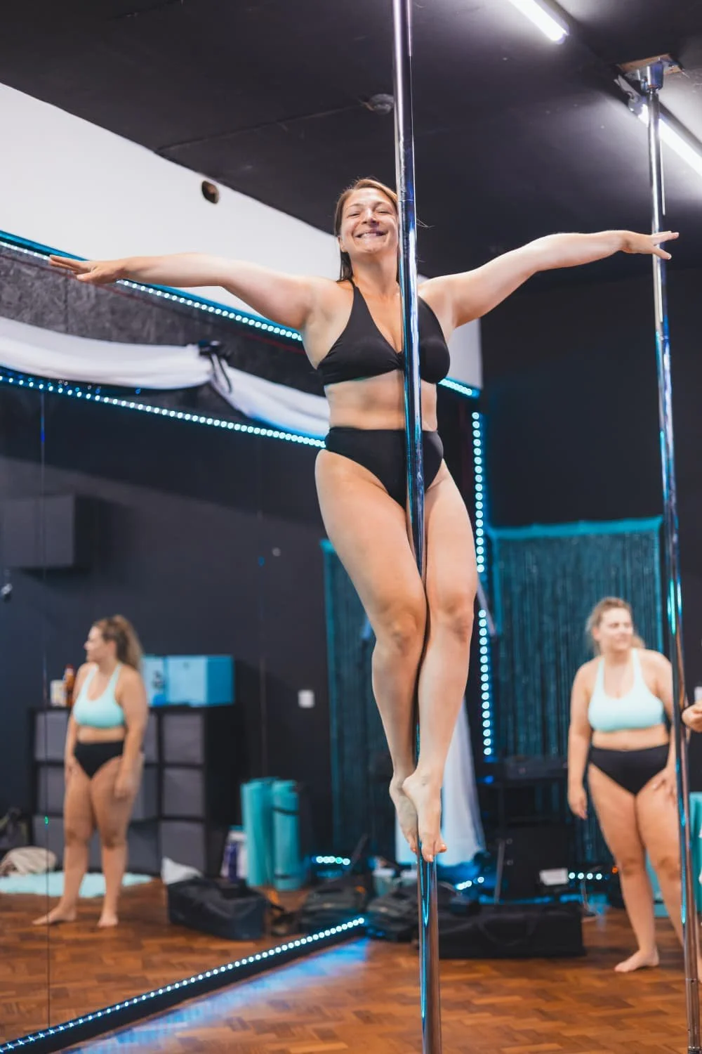 A woman practicing pole dance in a studio with black walls and mirrors, wearing black sports bra and shorts, smiling with arms outstretched.