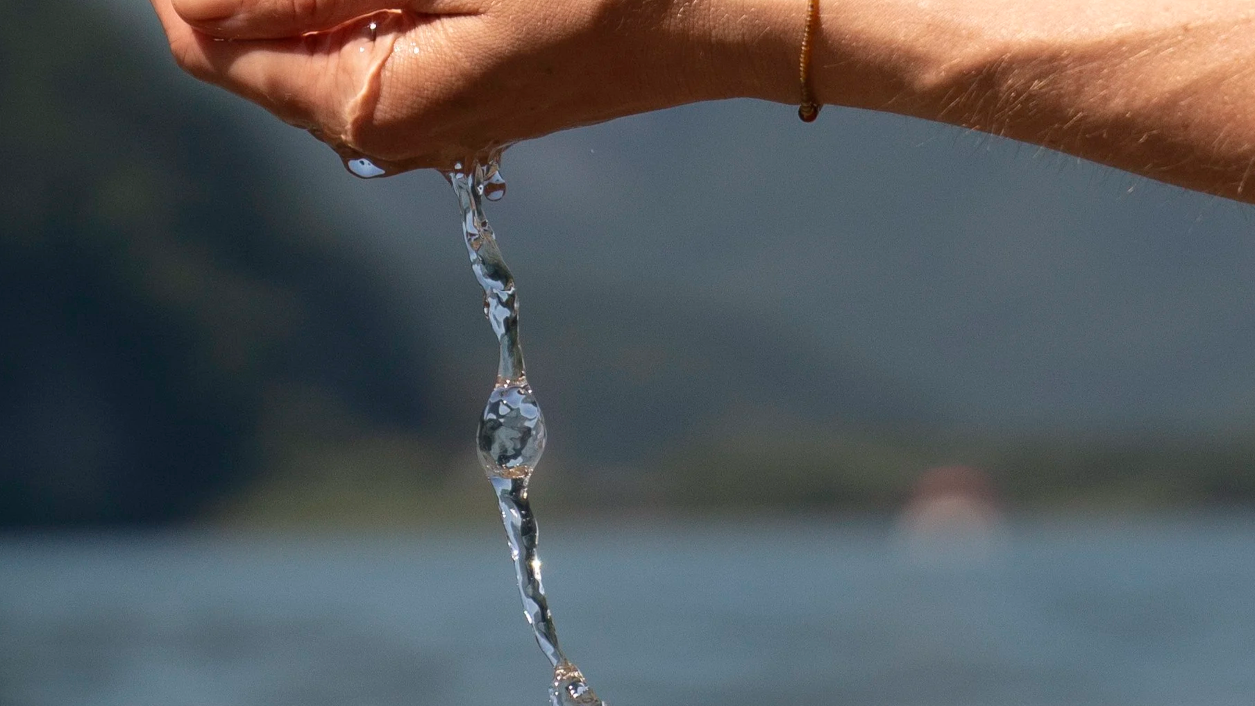 Hand hält Wasser, das in der Luft tropft, mit Wasser im Hintergrund. Naturheilkunde. Origin-Massage. SomabyJordan