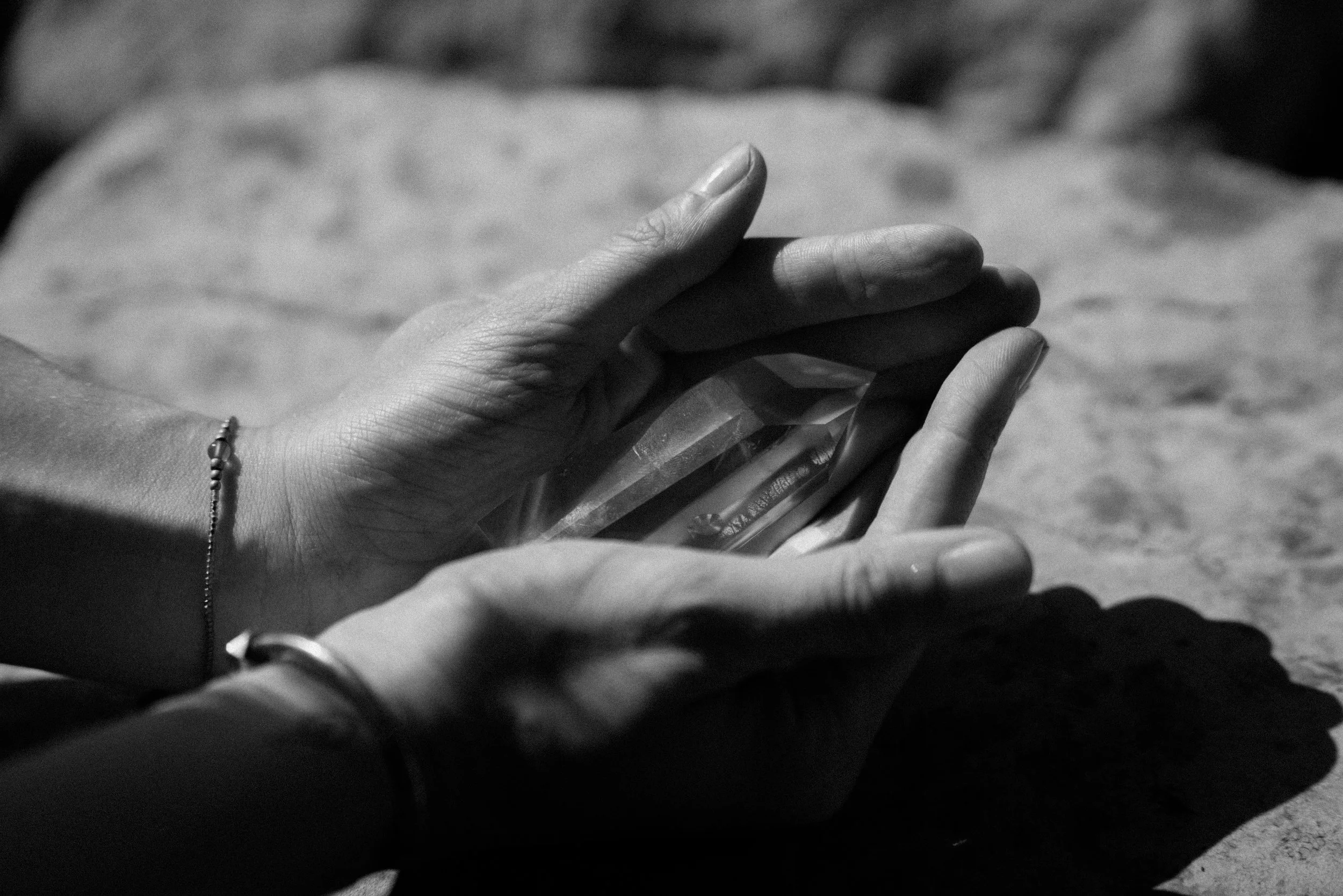 Schwarz-weiß-Fotografie von zwei Händen, die eine Kristall oder einen Edelstein halten, auf einem flachen Stein. Naturheilkunde. Origin-Massage. SomabyJordan
