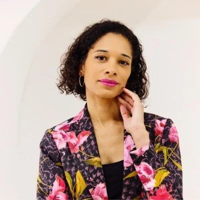 A woman with curly hair waving and smiling, wearing a black and white patterned sweater against a plain background.
