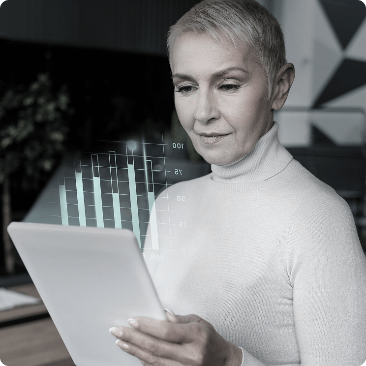 An older woman with short gray hair and a white turtleneck shirt is looking at a tablet. There is a digital overlay showing a bar graph and line chart in front of her, suggesting analysis or data presentation.