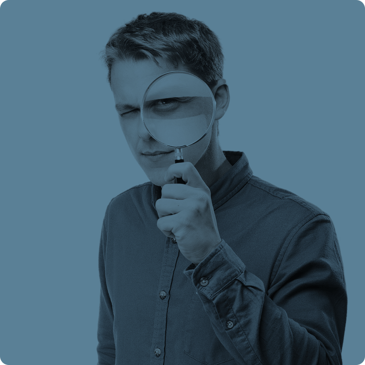 Young man holding magnifying glass up to his eye, winking, dressed in a button-up shirt, against a plain background.