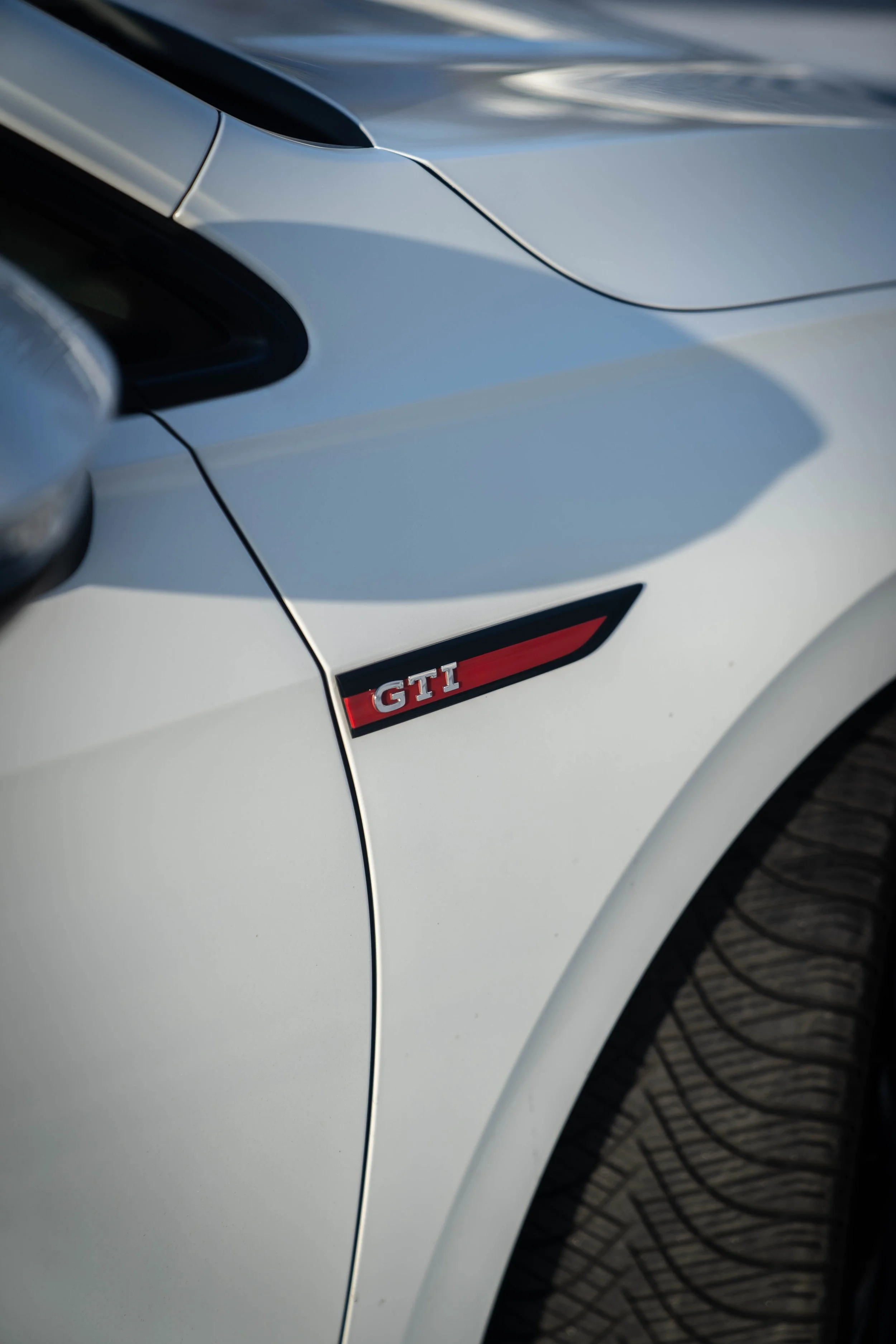 Close-up van een witte auto met een GTI badge aan de zijkant en delen van de koplamp en wiel zichtbaar.