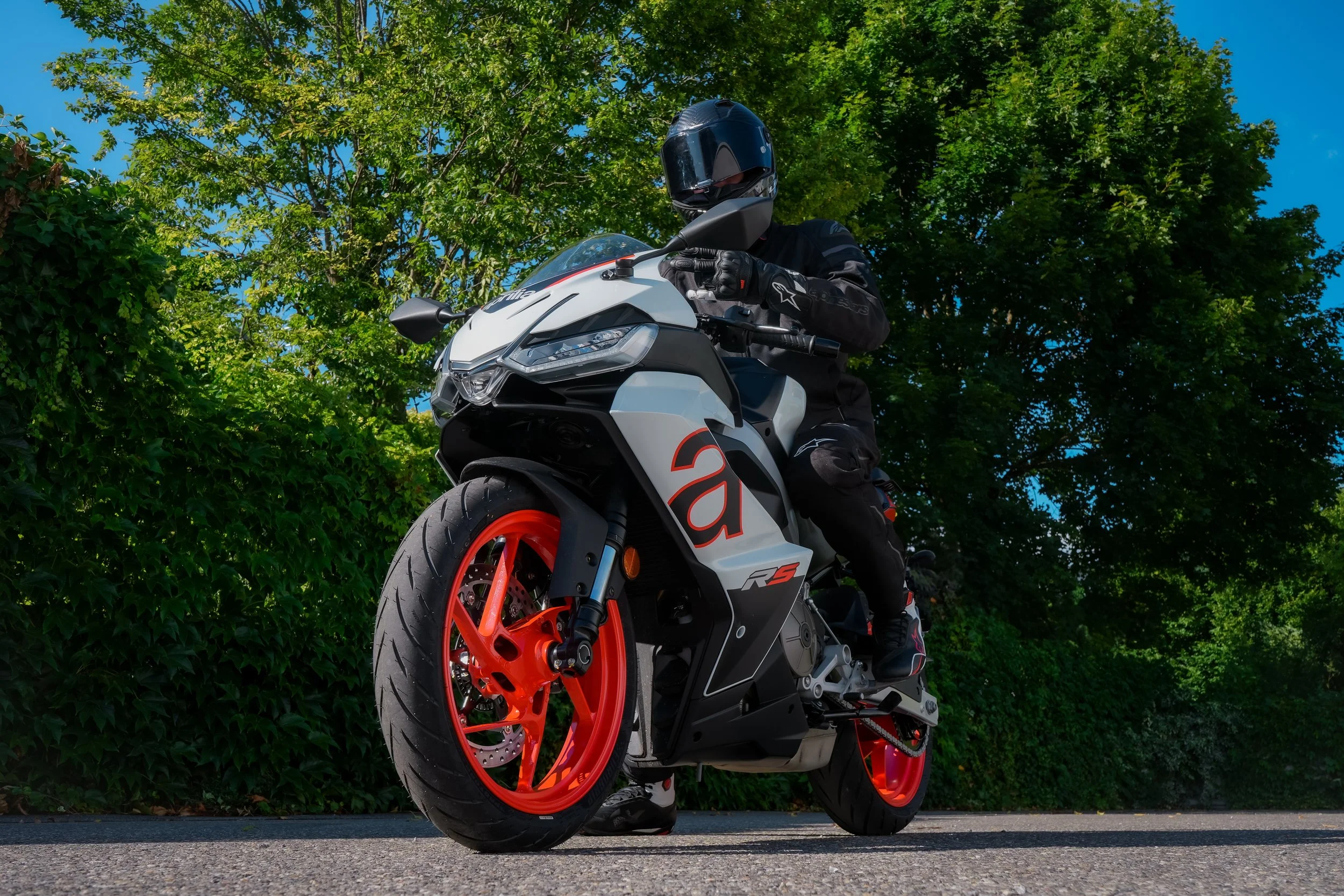 Motorradfahrer auf einer sportlichen weißen und schwarzen Rennmaschine mit roten Felgen, getragen von einem schwarzen Helm und schwarzer Schutzkleidung, auf einer Straße mit grünen Büschen im Hintergrund.