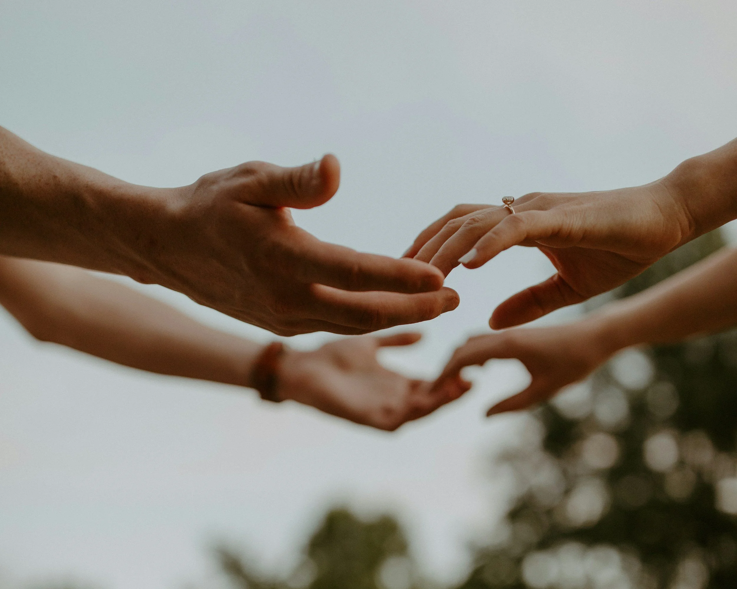 Two hands reaching out to each other, one with a ring on the finger, with a blurred outdoor background.
