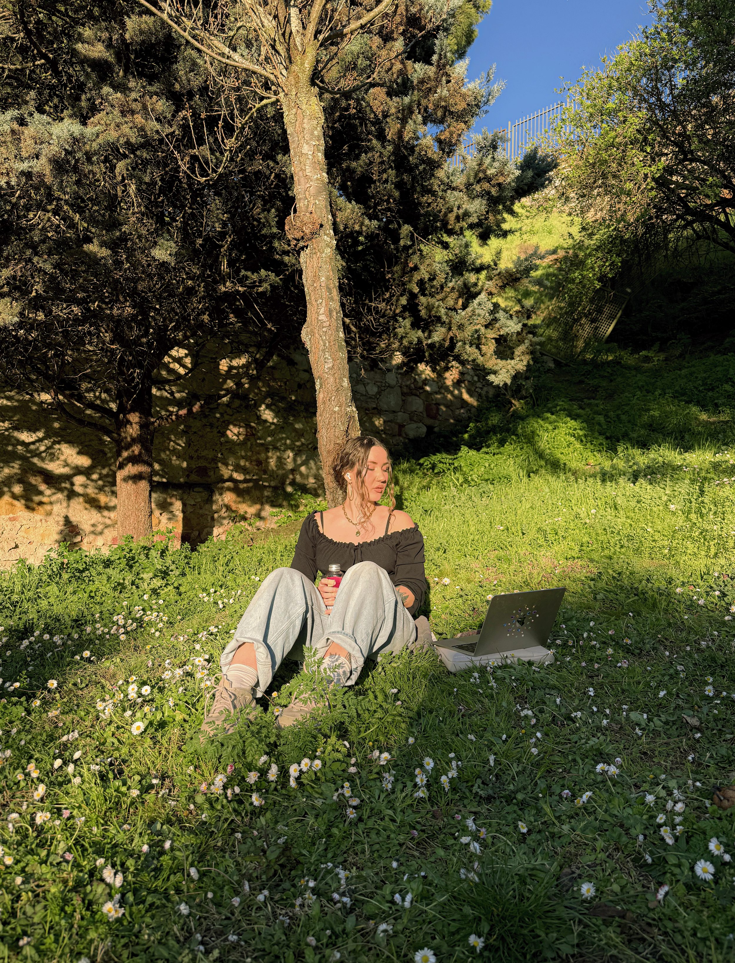 A young woman with long brown hair sitting on the grass under a tree, holding a bottle and looking at her laptop, with small white flowers and green foliage around her.